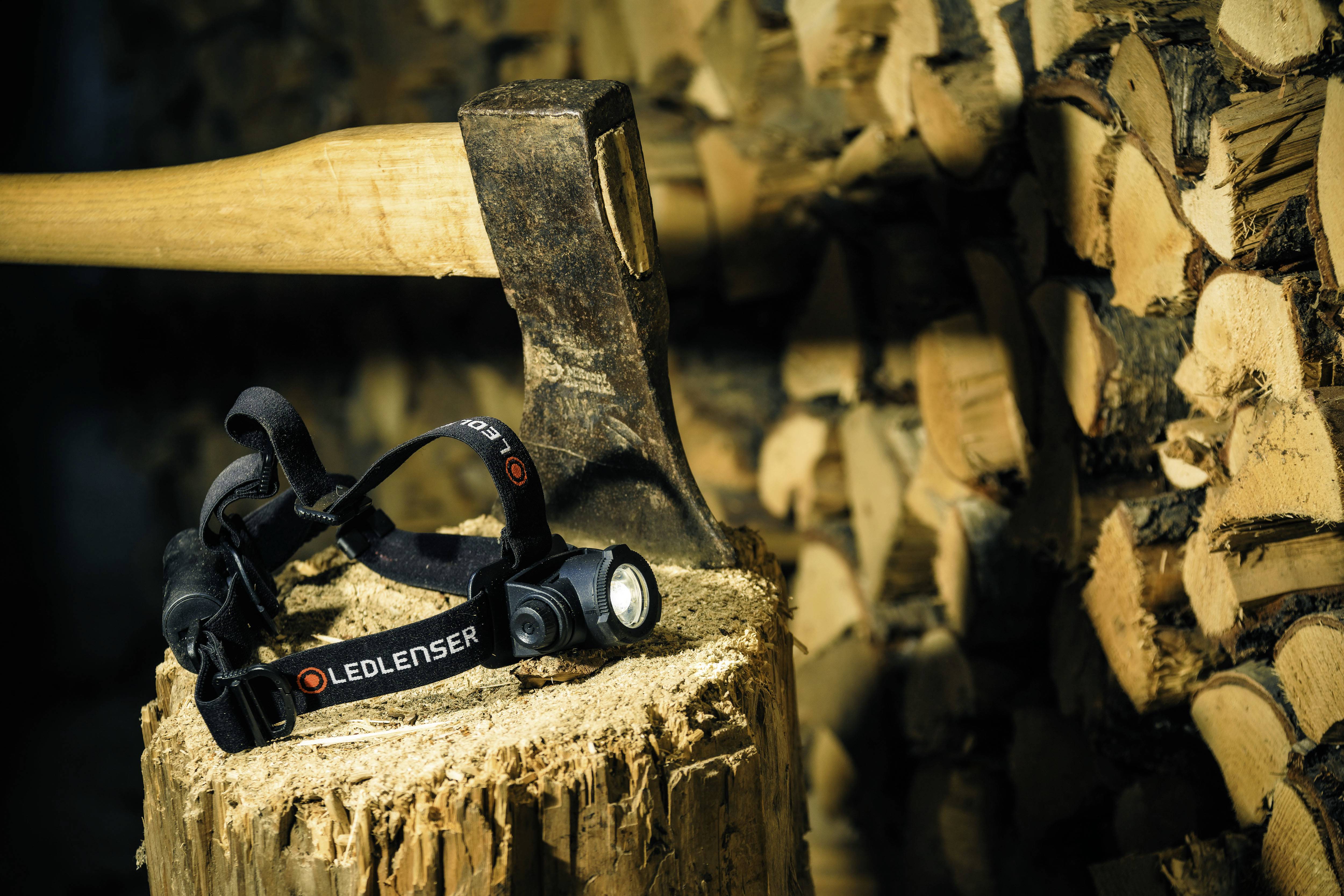 An axe on a tree stump with an LED head torch beside it; background with stacked firewood.