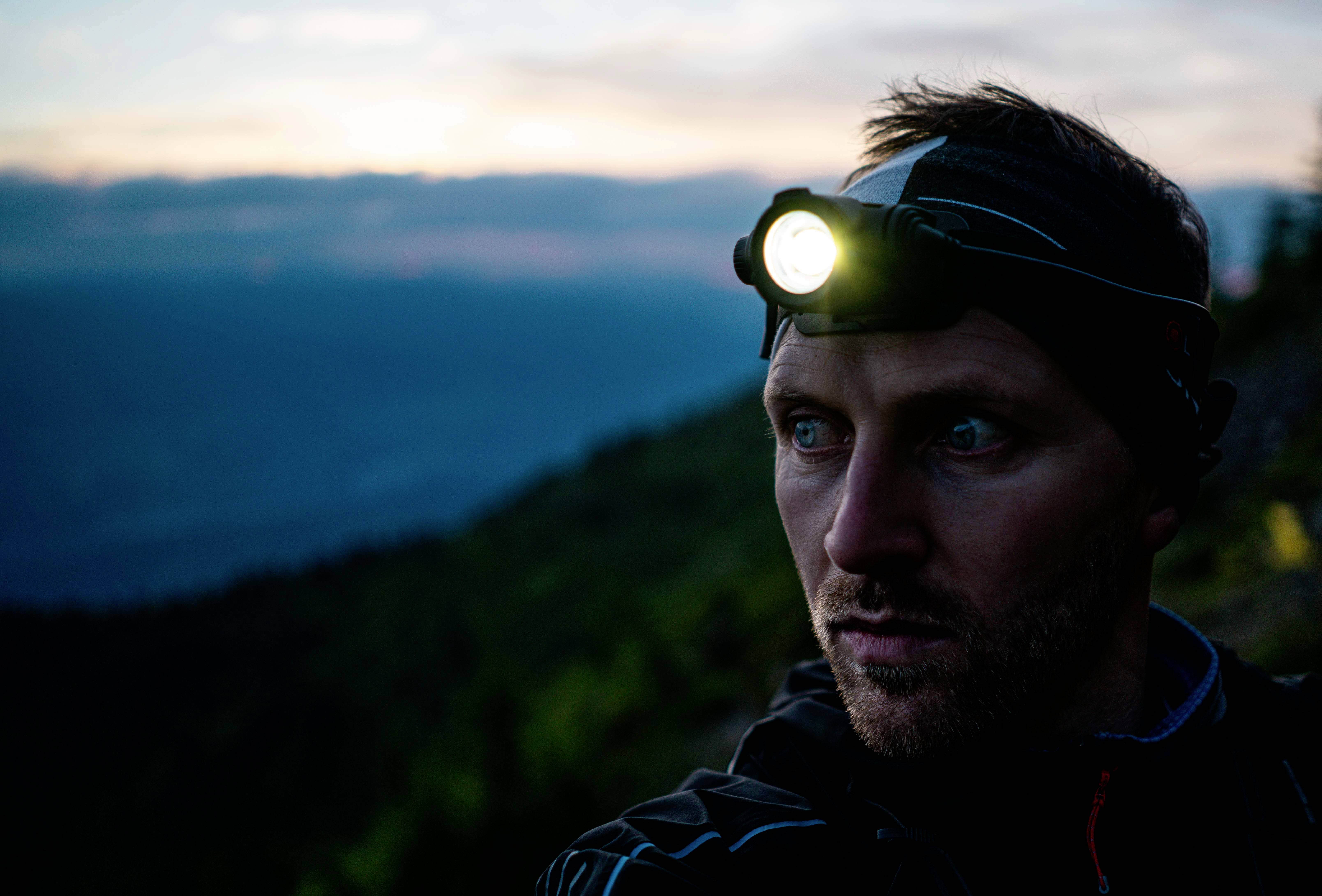 A man wearing a head torch gazes into the distance, with a landscape at dusk in the background.