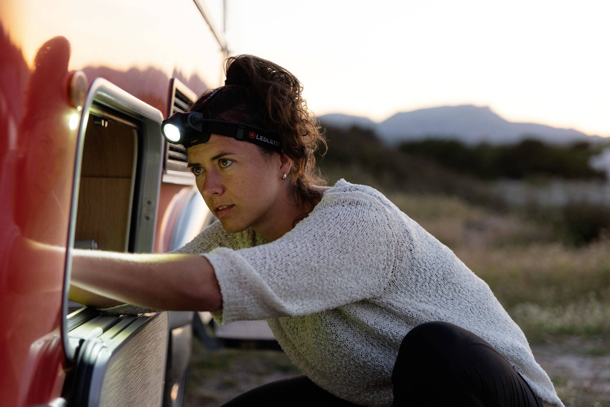 A person wearing a head torch is working intently on a red vehicle in a rural setting at dusk.