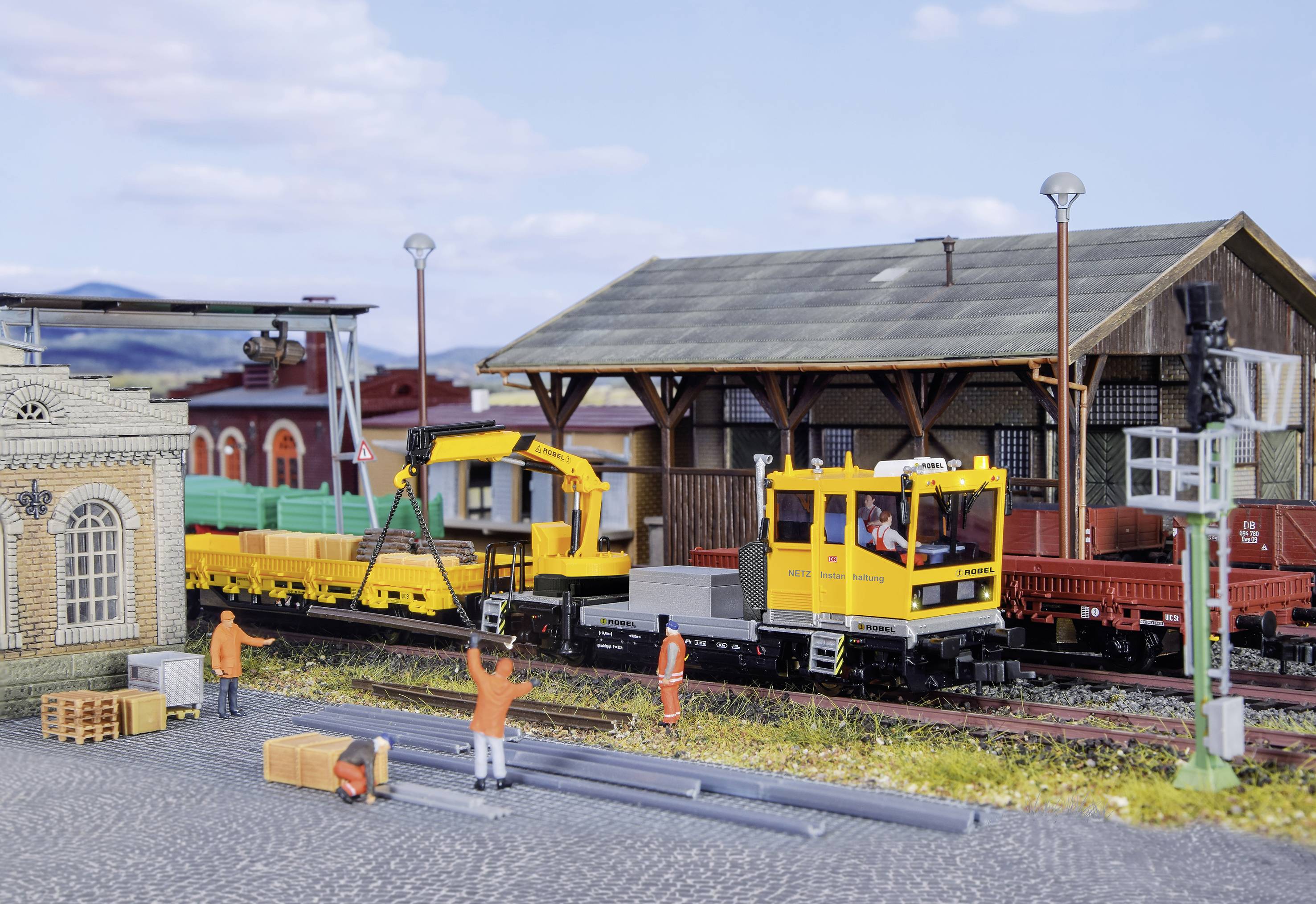 A scene at a model railway station: Workers are loading wagons with pipes and crates. A yellow crane and wagons are standing ready.