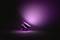 A modern table lamp lying on its side, casting violet light behind it onto a black surface, creates atmospheric lighting.