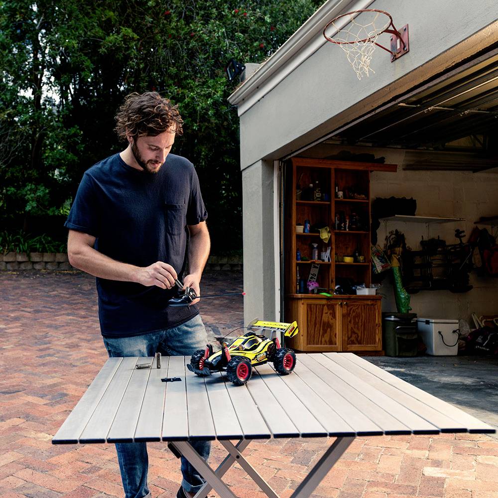 A man is controlling a remote-controlled car on a table in front of an open garage. Enthusiastic play outdoors.