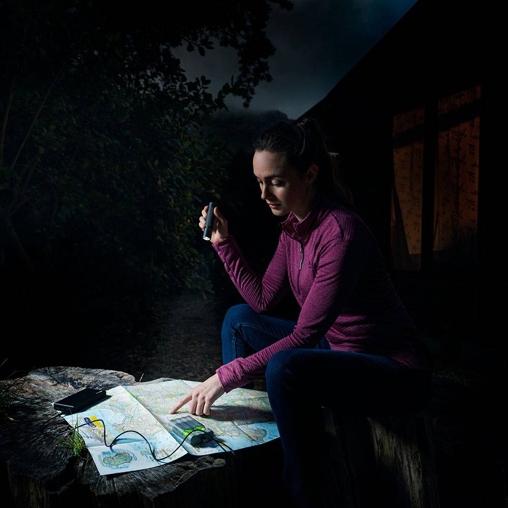 A woman sits outdoors, holding a smartphone and studying a map on a table, illuminated by the phone's screen.