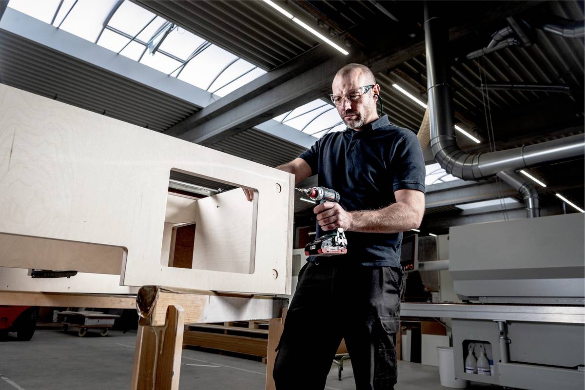 A man in a workshop is drilling a large rectangular hole in a wooden board. Daylight is streaming through the roof window.