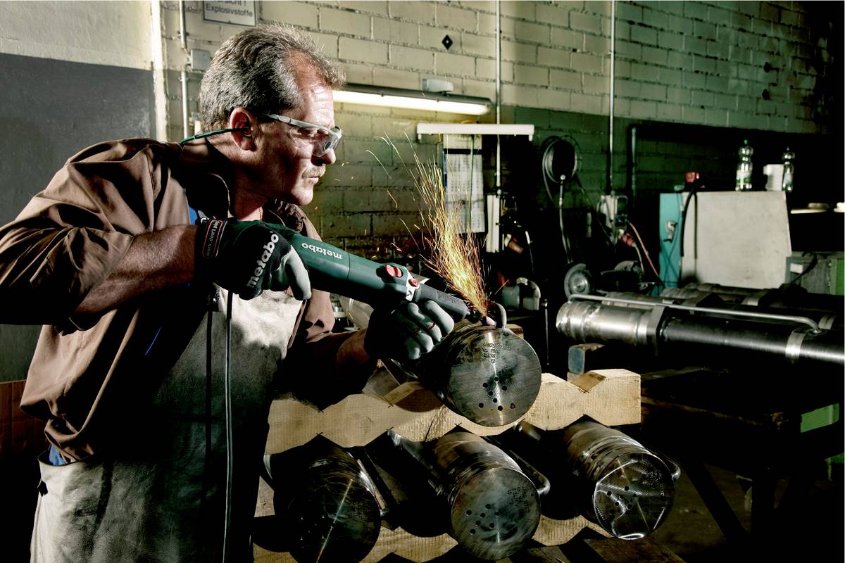 A man in a workshop is using an angle grinder to work on metal pipes. Sparks are flying from the grinding point.