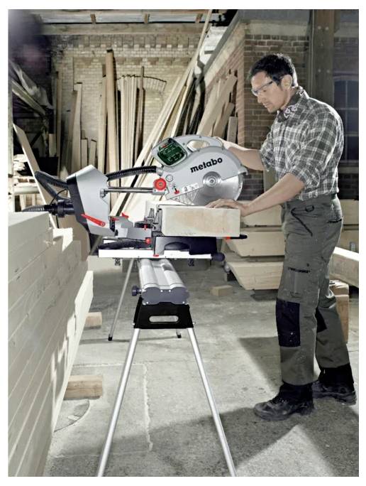 A man uses a Metabo miter saw to cut a wooden plank in a workshop filled with lumber.
