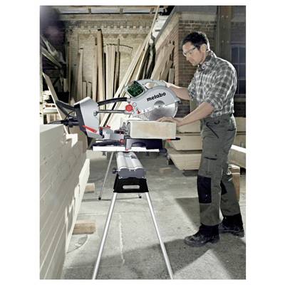 A man uses a Metabo miter saw to cut a wooden plank in a workshop filled with lumber.