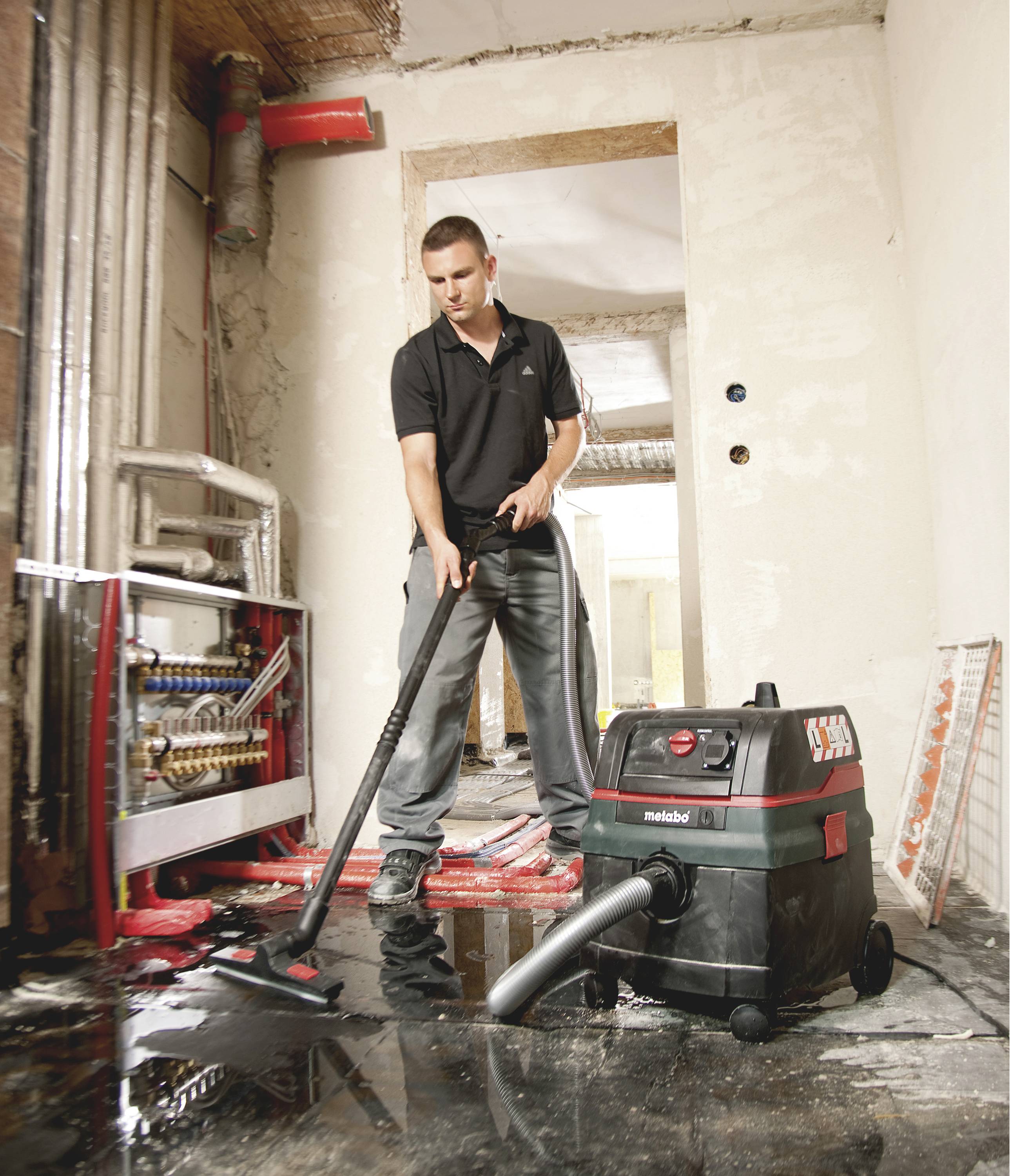 A person cleans a flooded room using an industrial vacuum, surrounded by construction materials and pipes, suggesting renovation work.