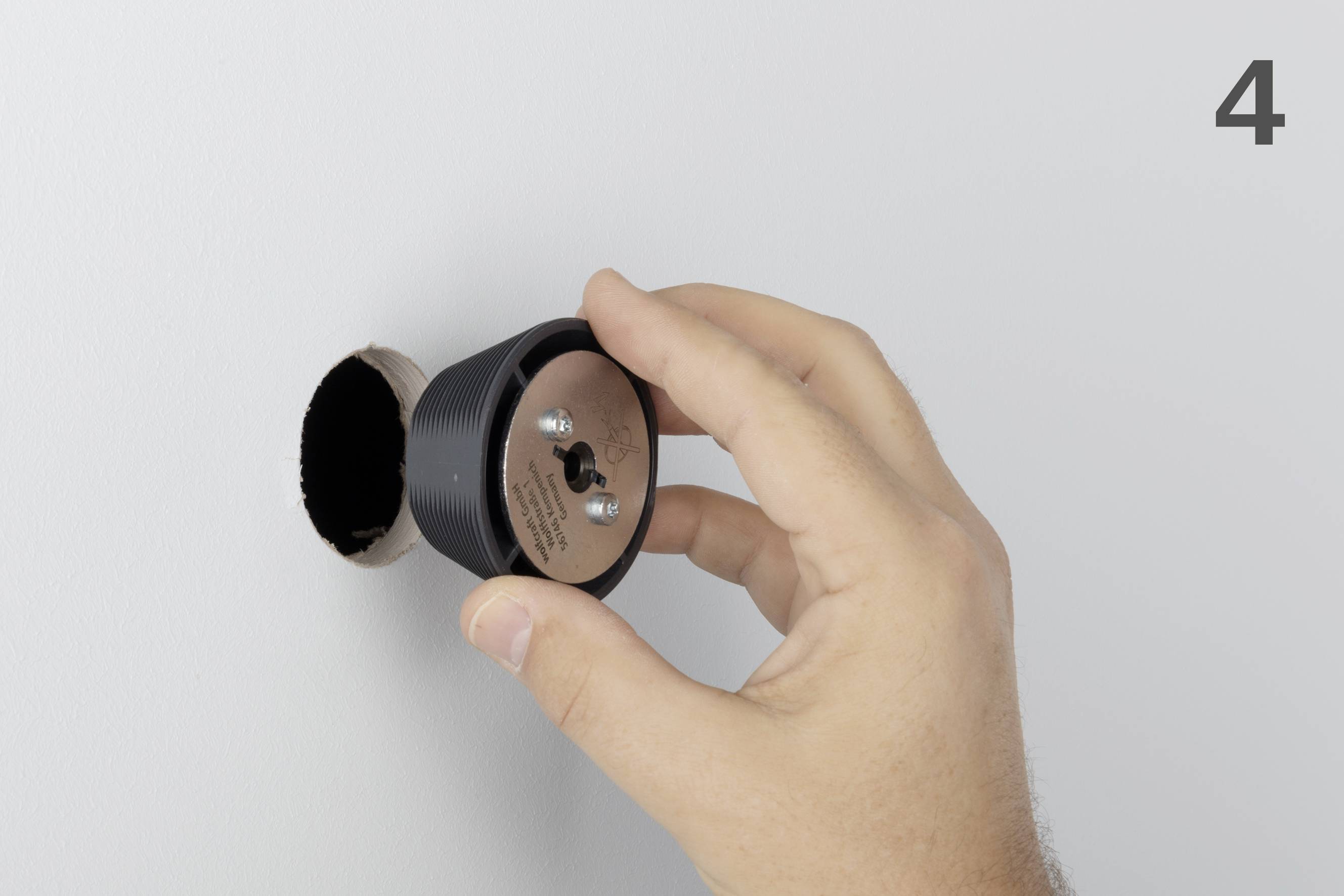 A hand is inserting a black wall mount into a circular hole in a white wall.