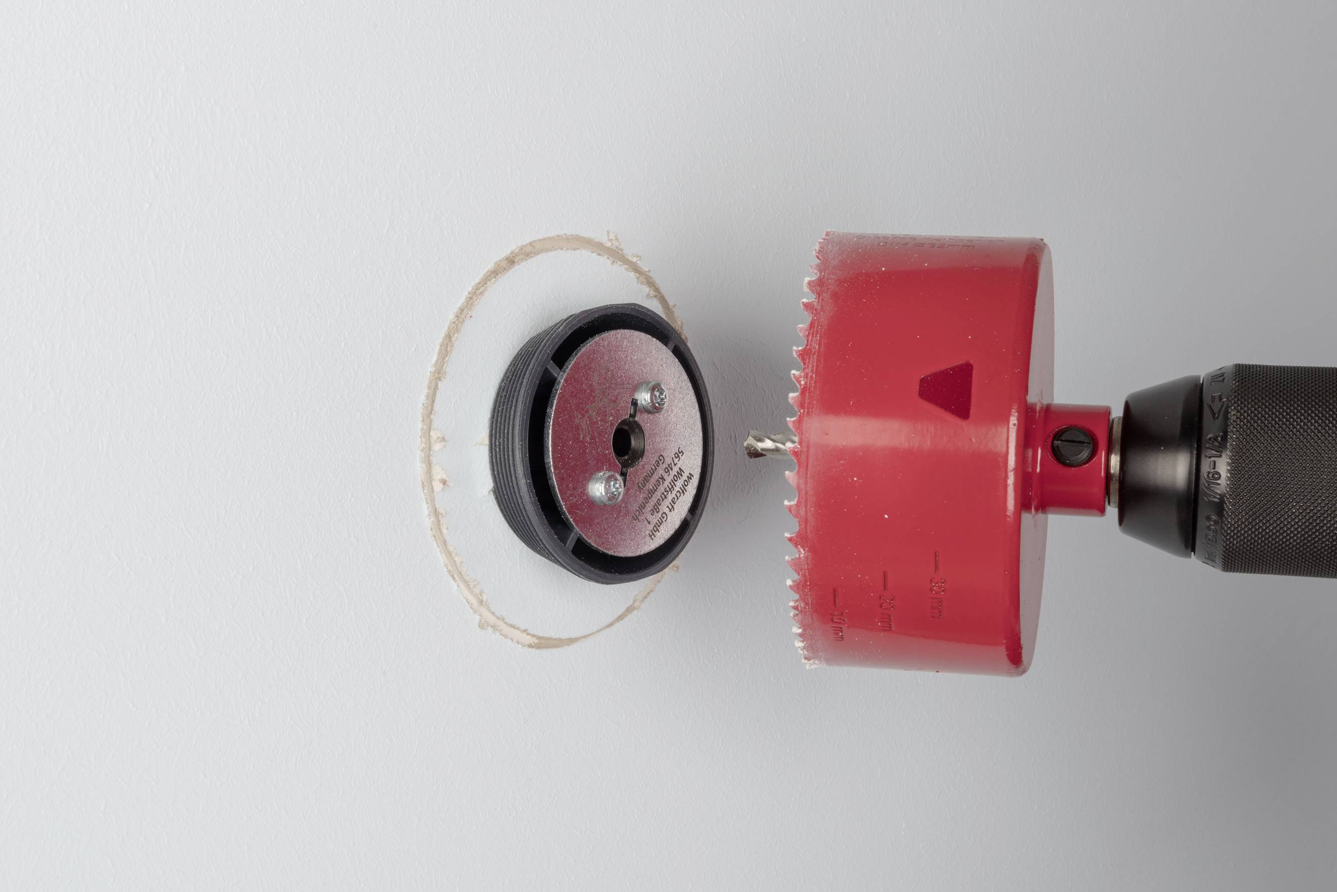 A tool with a red drill bit creates a round hole in a light-coloured wall. The drill bit is shown in close-up.