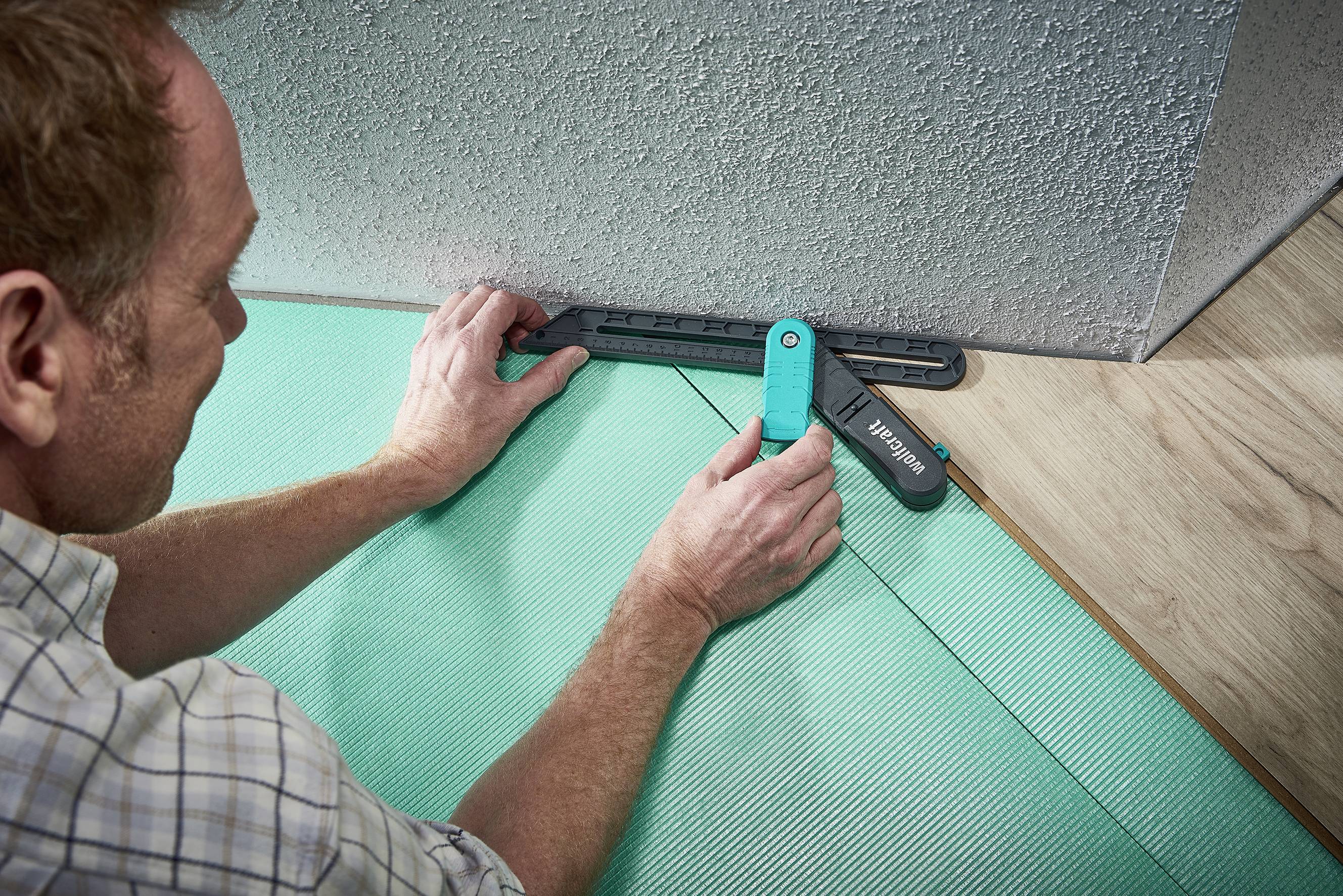 A man is measuring the edge alignment of a floor against a wall using a measuring tool. He is preparing the surface for floor installation.