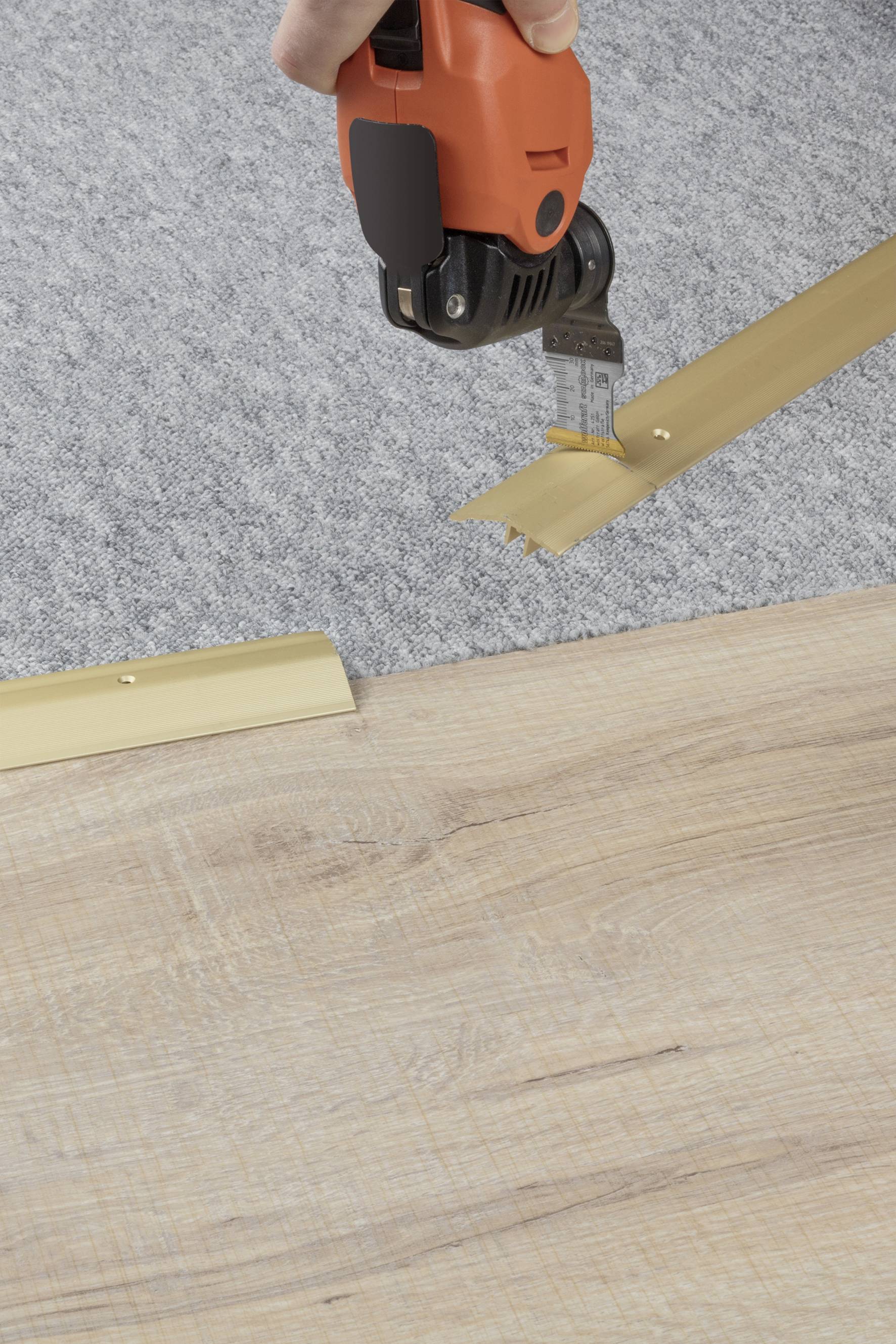 A person is using an electric cutting tool to cut a gold-coloured metal profile on a wooden and carpeted floor.