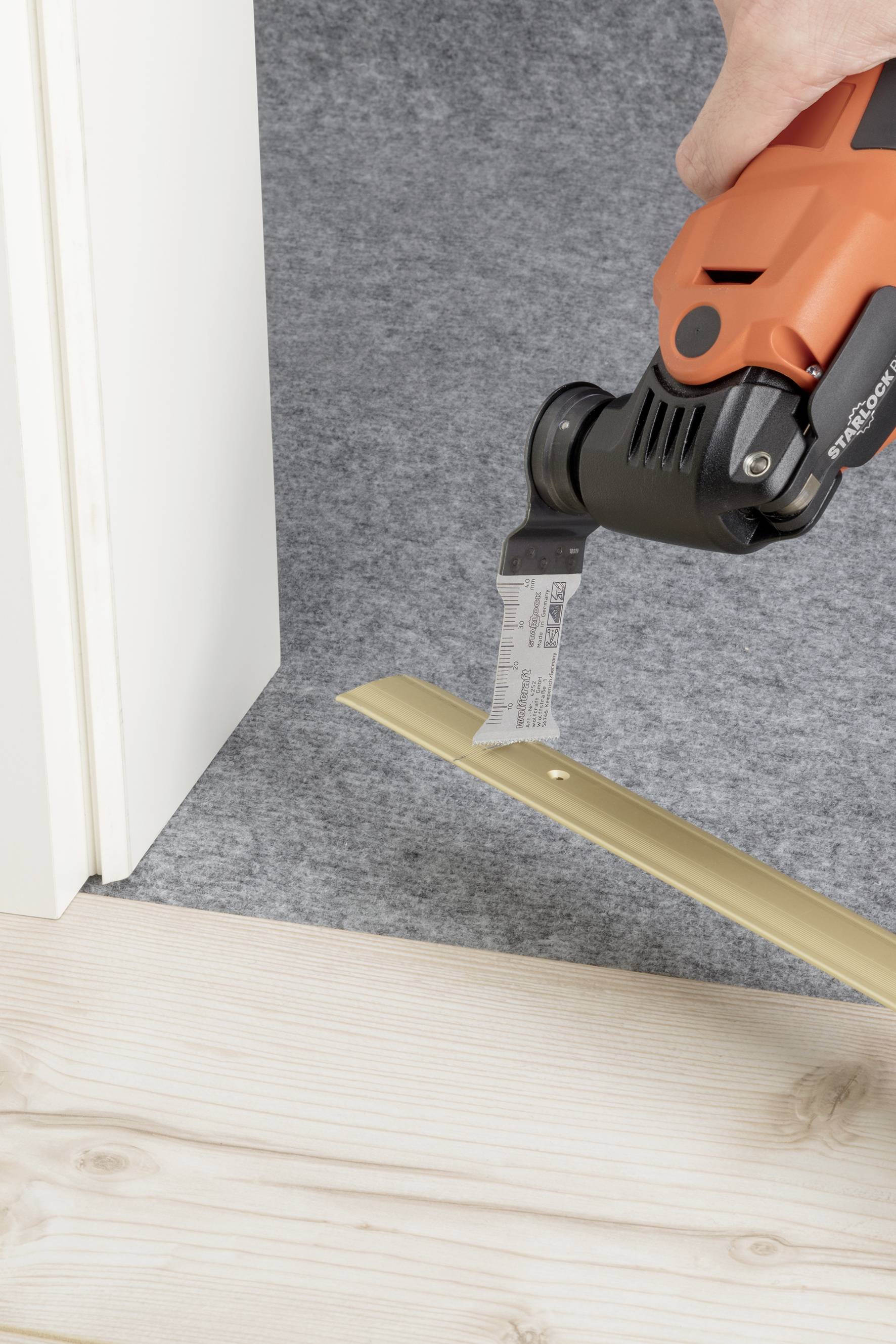 A person is cutting a golden metal rail with an oscillating saw on a wooden floor next to a door frame.