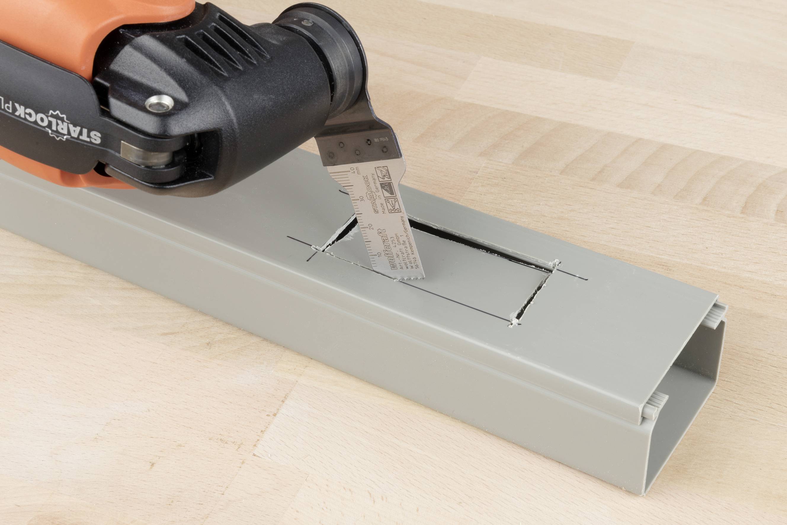 An oscillating tool with a saw blade is cutting a rectangular cut-out in a grey plastic sheet.