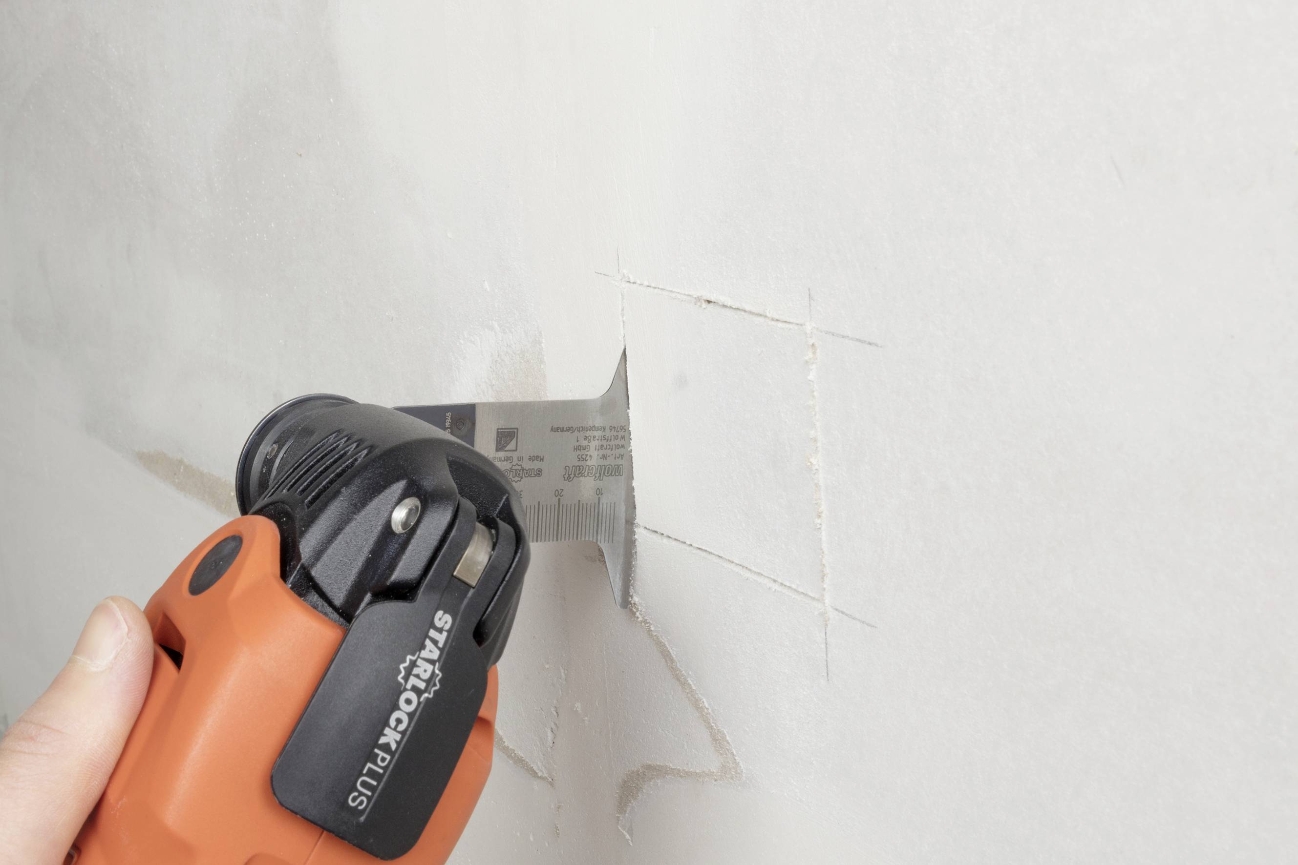 A person is cutting a square hole in a plasterboard wall using an orange electric tool.