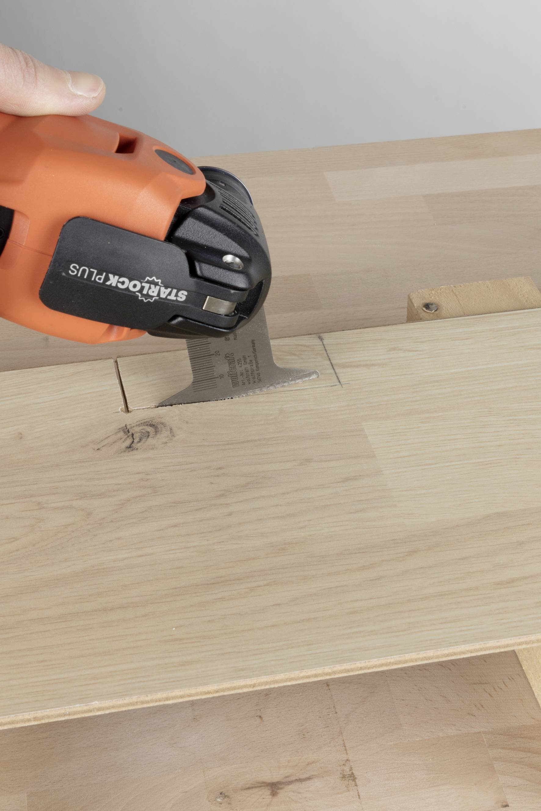 A hand is holding an orange tool cutting into a wooden board to perform precise repairs or adjustments.