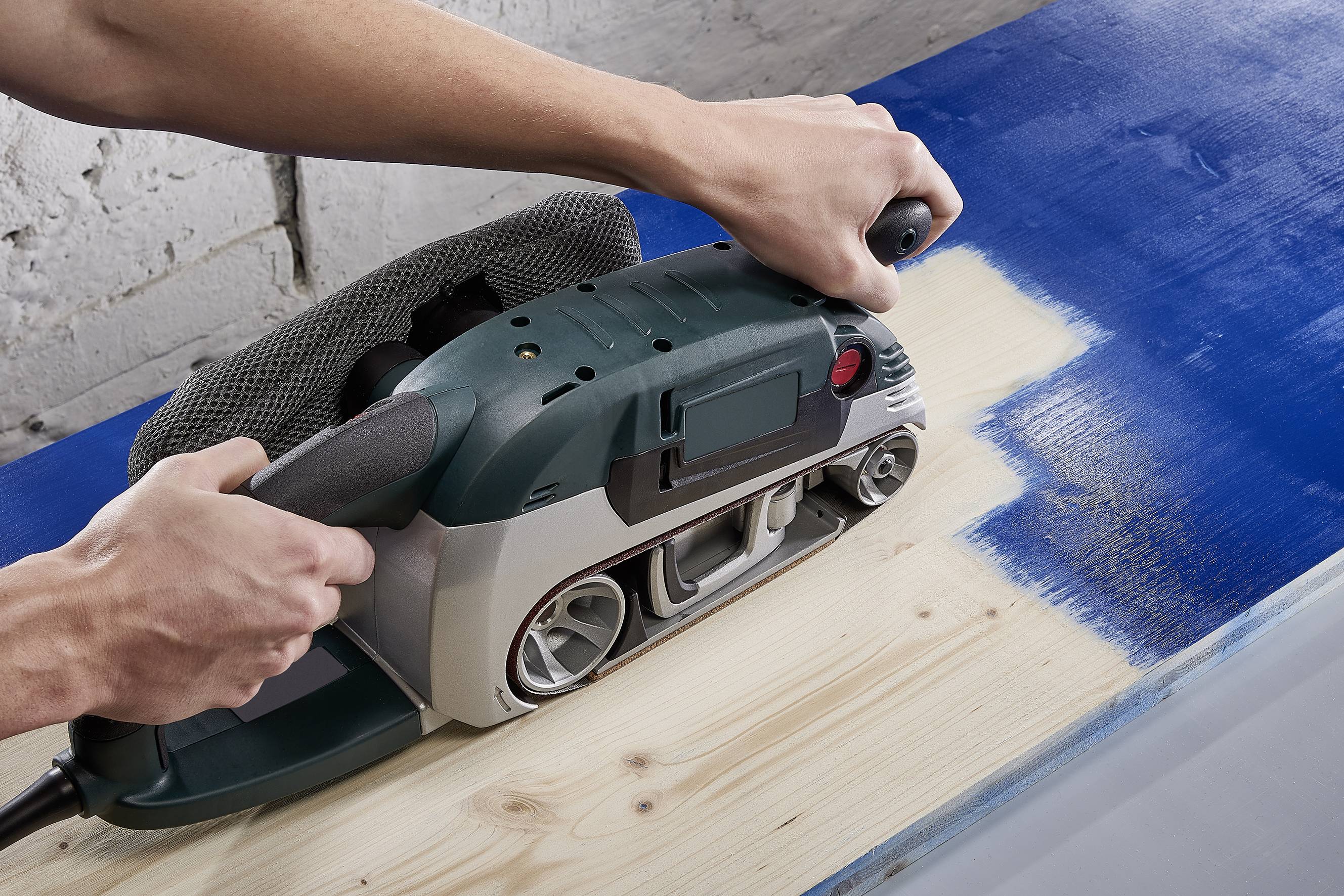 A person is sanding blue wood with a belt sander to smooth it by removing the coloured layer.