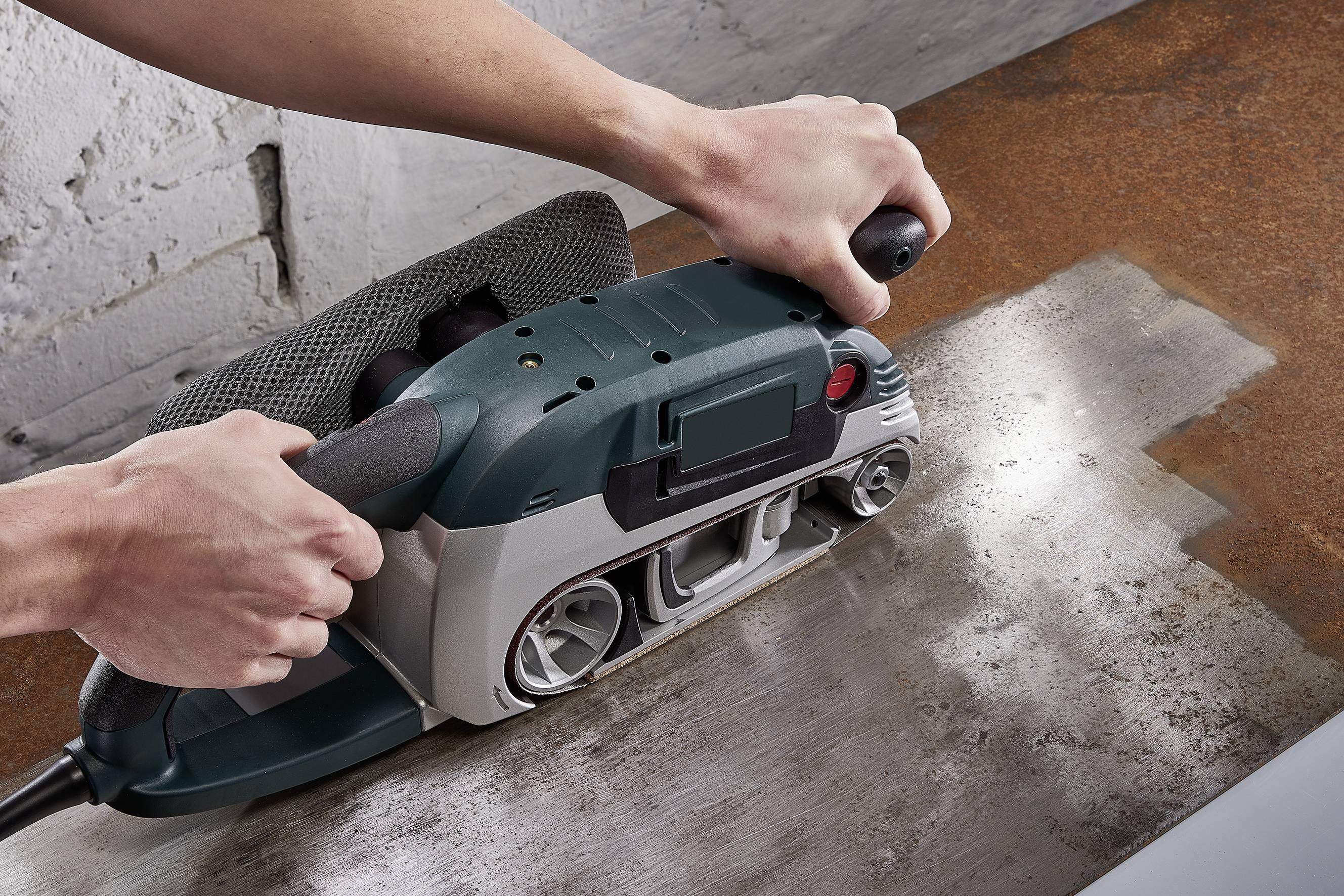 A person is sanding a metal surface with an electric belt sander to remove rust and smooth the surface.