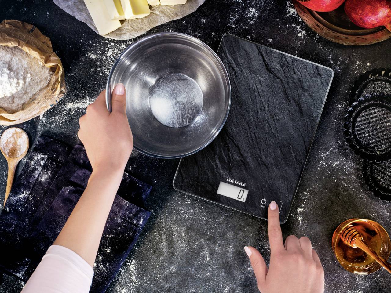 A hand holds a metal bowl on a digital kitchen scales, surrounded by baking ingredients on a table.