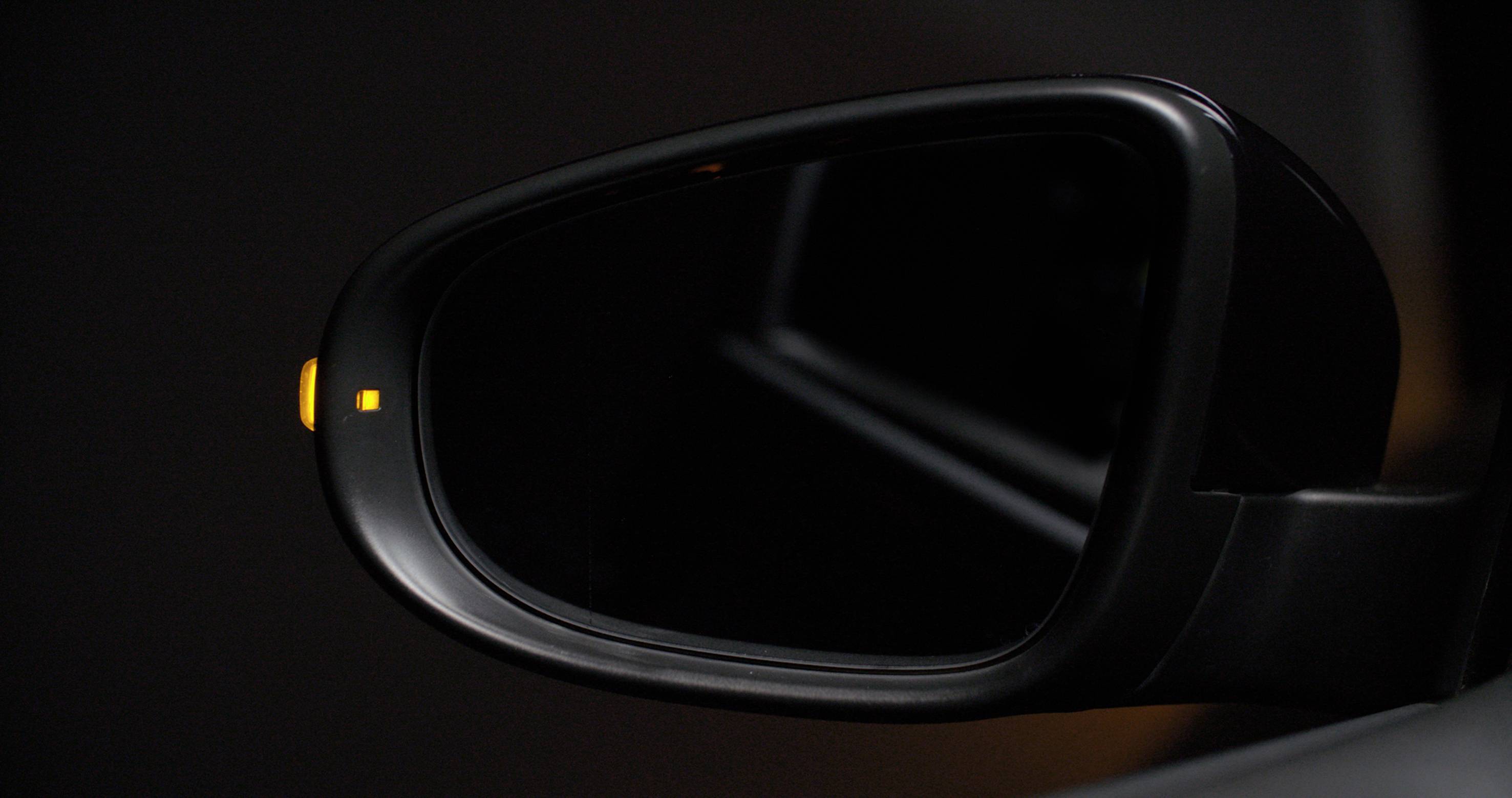A car rear-view mirror with a yellow warning symbol indicates the presence of a vehicle in the blind spot.