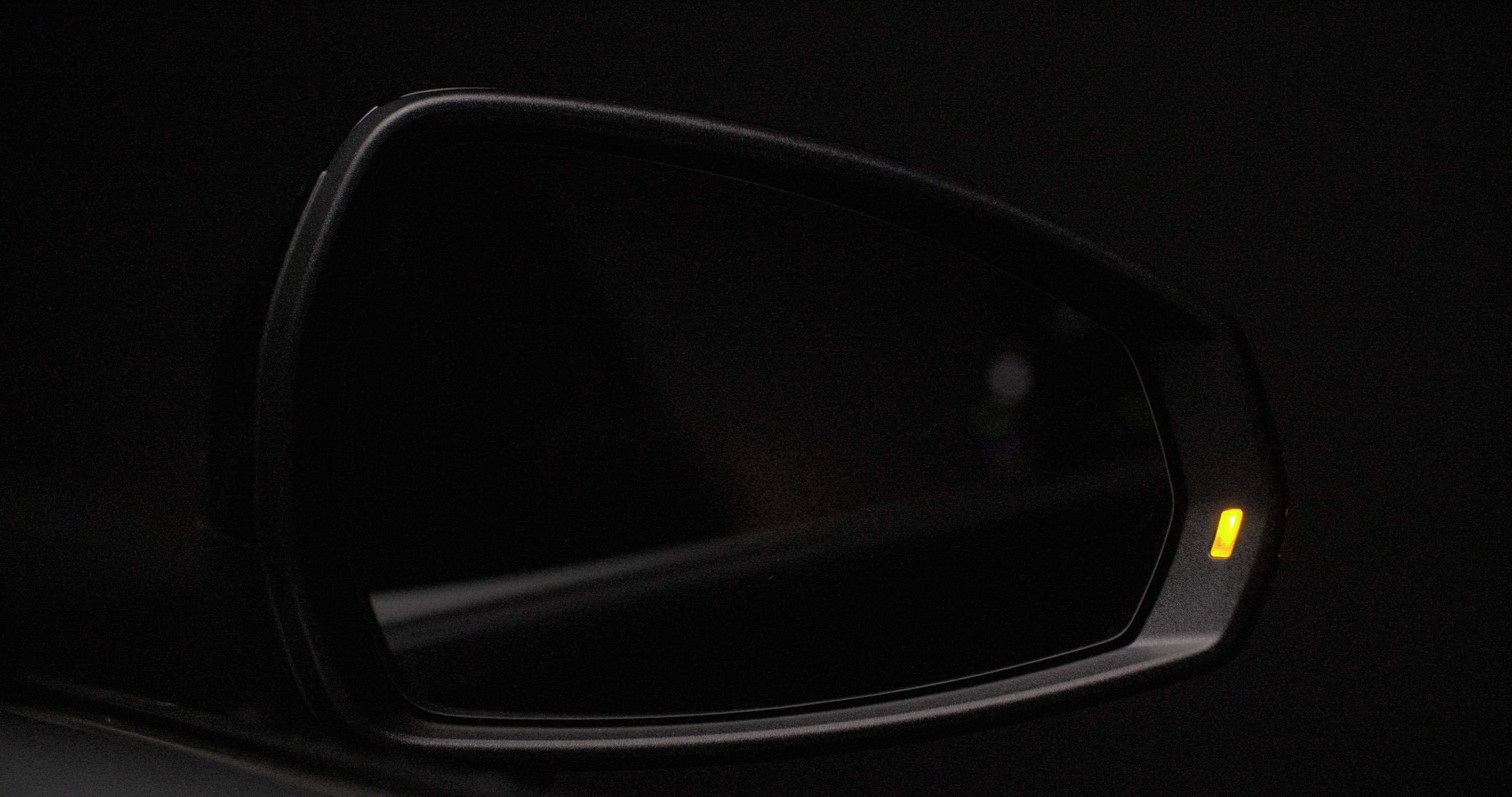 A dark car wing mirror with an active yellow warning light, signalling approaching traffic in the blind spot.