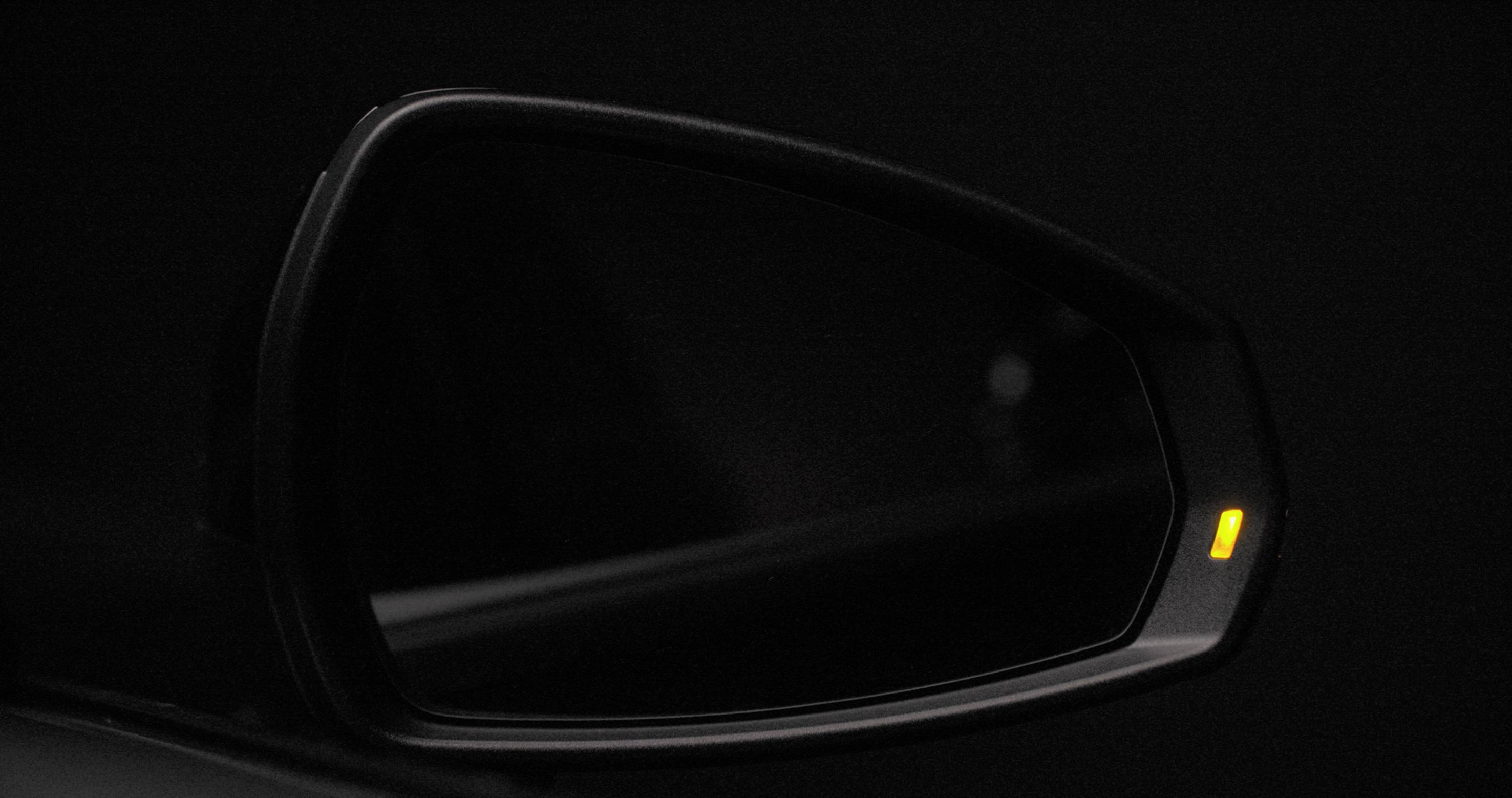 A vehicle wing mirror with a small yellow light, likely indicating a blind spot, against a dark background.