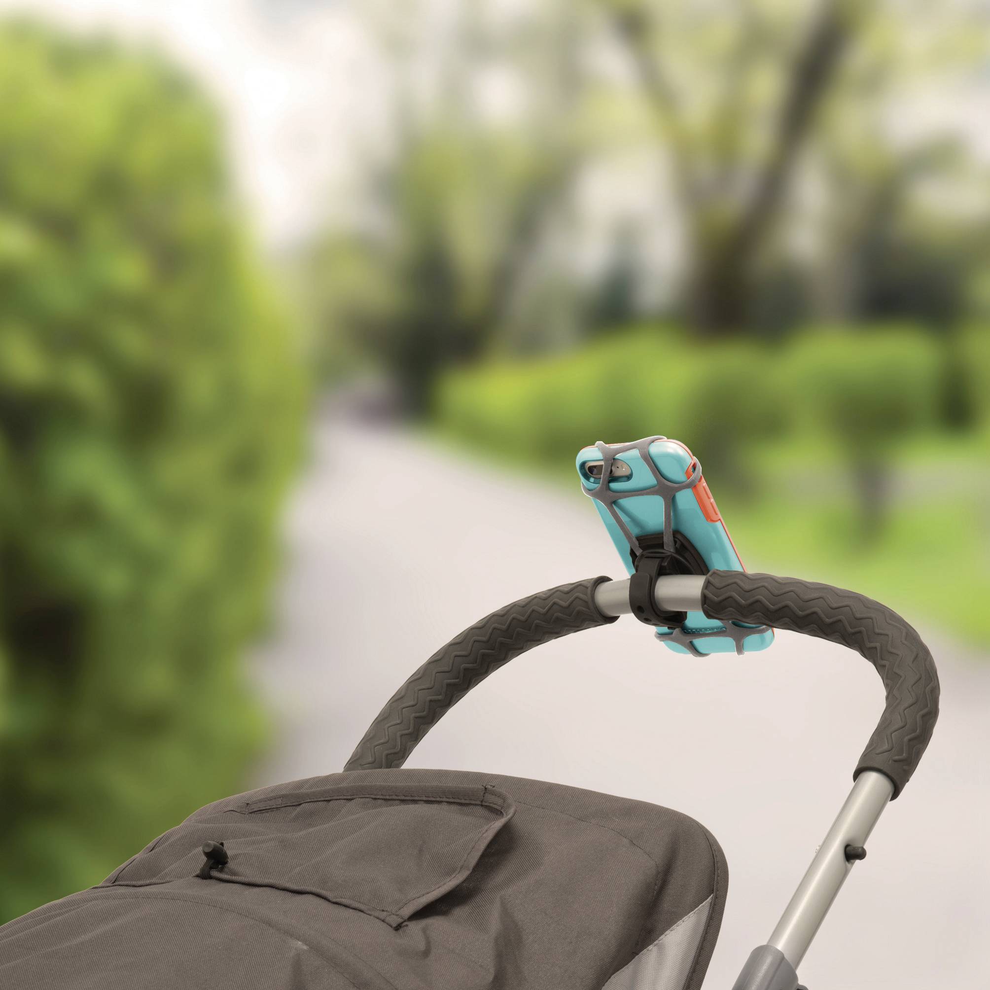 Pushchair on a paved path, with a smartphone attached to the handlebar. Background blurred, with green bushes.