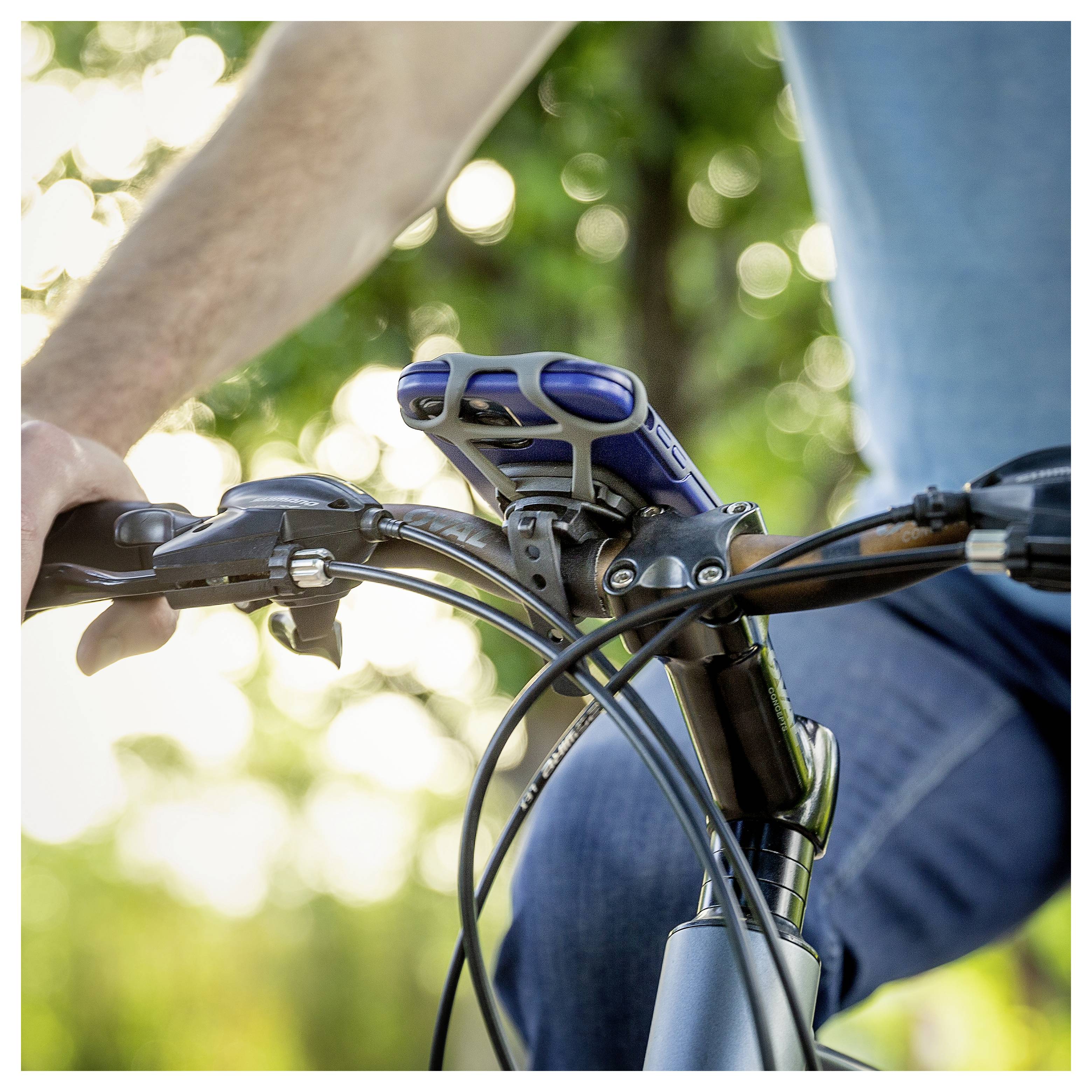 A person rides a bicycle, holding the handlebars with a smartphone mounted at the center. The background is blurred with greenery.