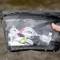 A hand holds a waterproof, transparent bag containing earphones and a cable, close to a body of water.