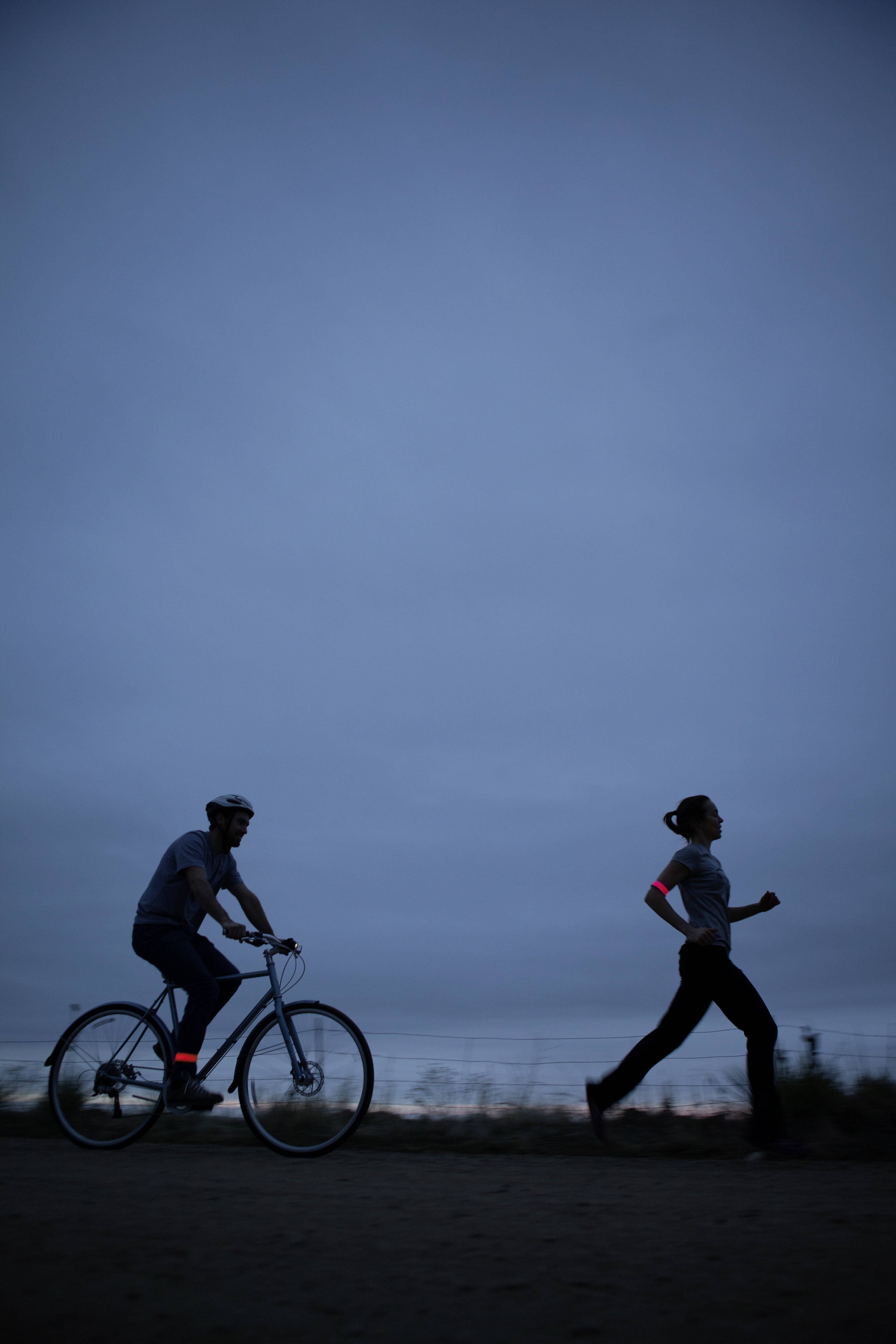 A person is jogging, another is cycling, both wearing reflective bands at dusk.