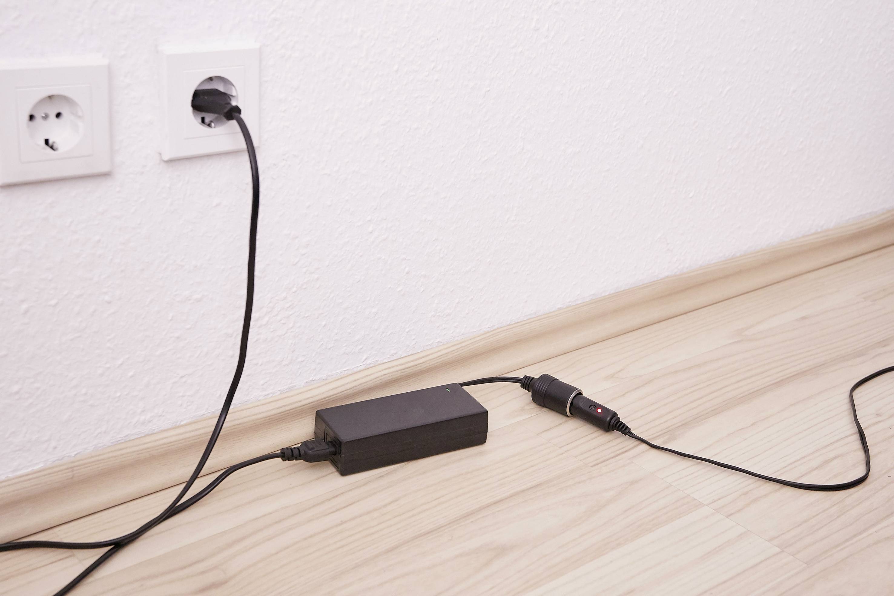 A black charger is plugged into a socket on a white wall and is resting on a light wooden floor.