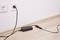 A black charger is plugged into a socket on a white wall and is resting on a light wooden floor.