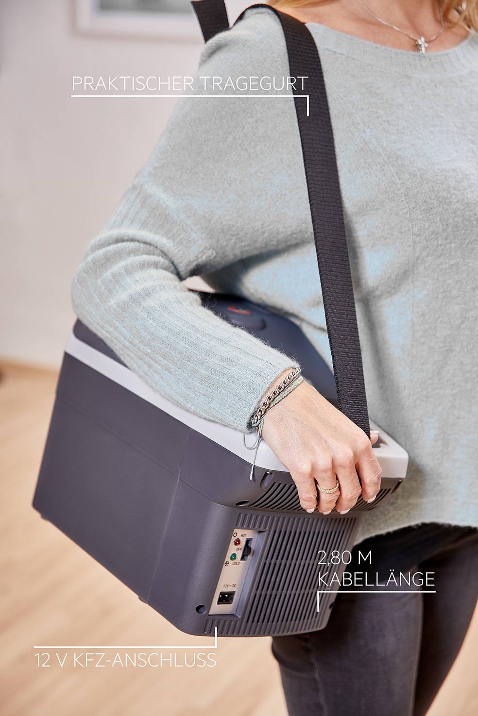 A person is carrying a portable cool box with a shoulder strap. The box has a 2.8 m long cable and a 12 V car socket.