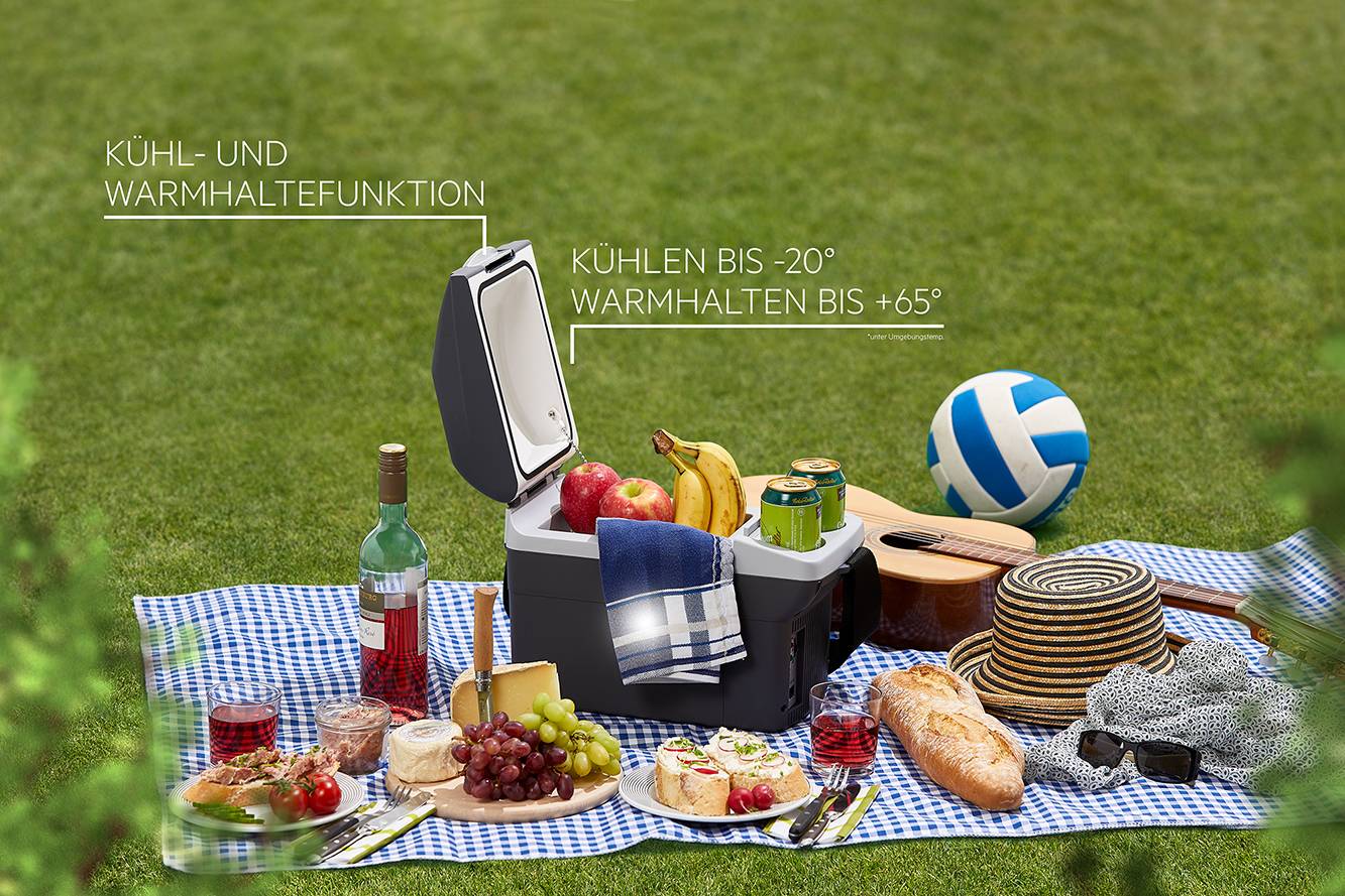 A picnic on a blanket in the countryside with a cool box, drinks, fruit, bread and crockery. A volleyball rests on the grass.