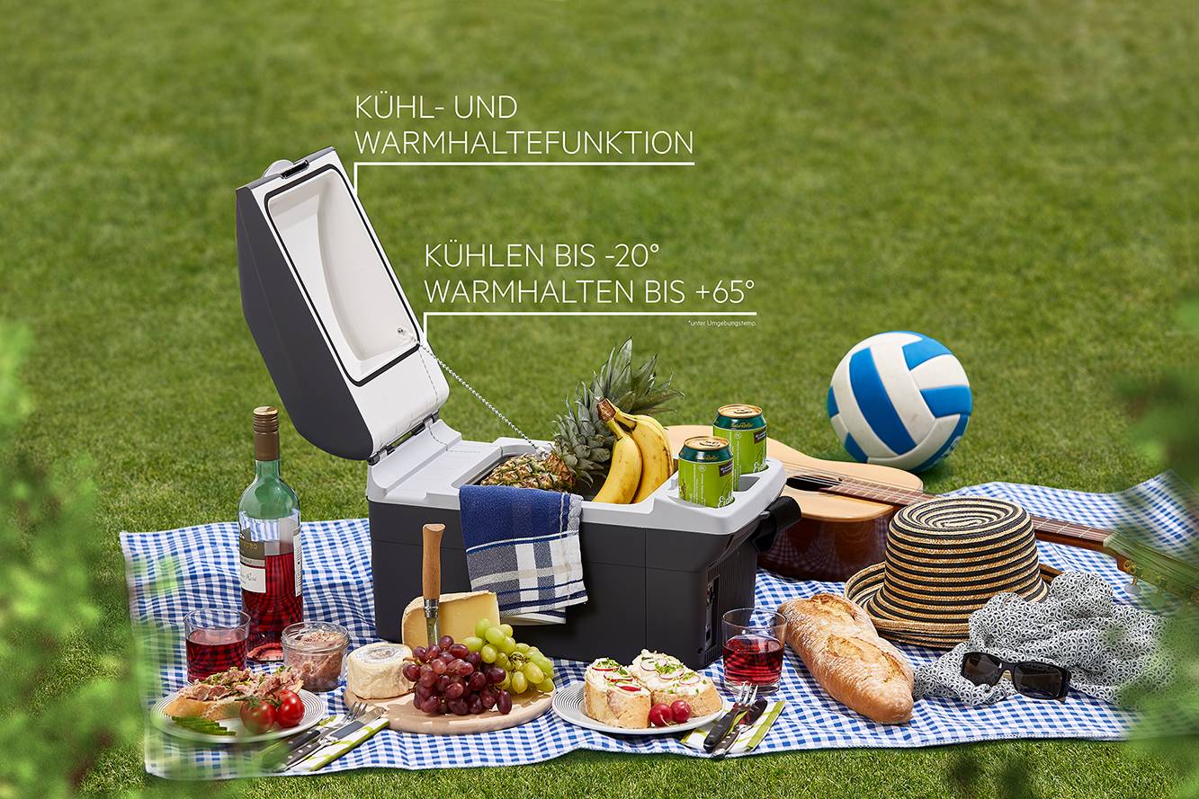 Picnic on a meadow: Cool box with open lid, food and drinks on a blanket, volleyball in the background. Text: 'Cool down to -20°, Keep warm up to +65°'.