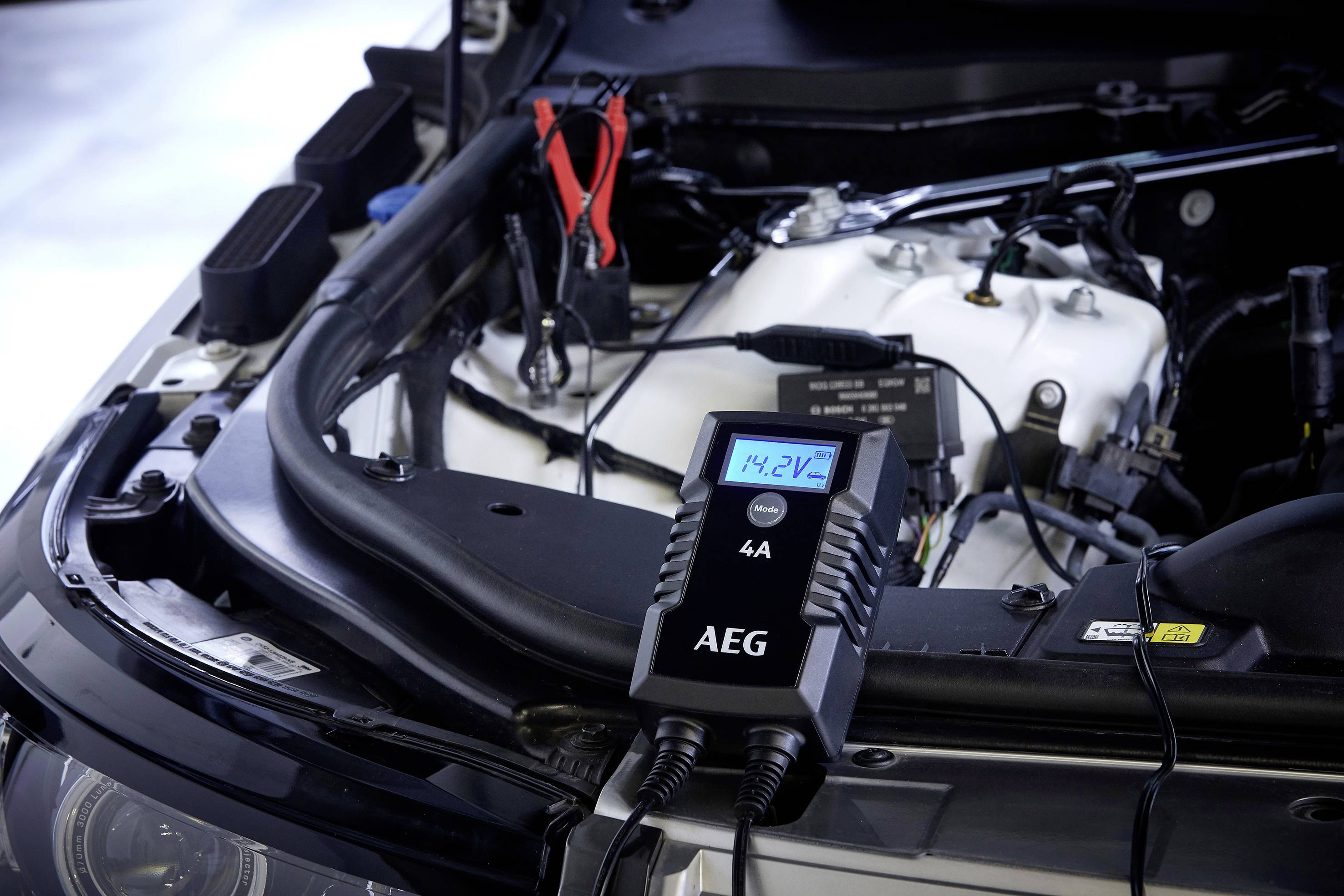 A car charger is connected to a car battery and displays '14.2V' on a display. Battery terminals are visible.