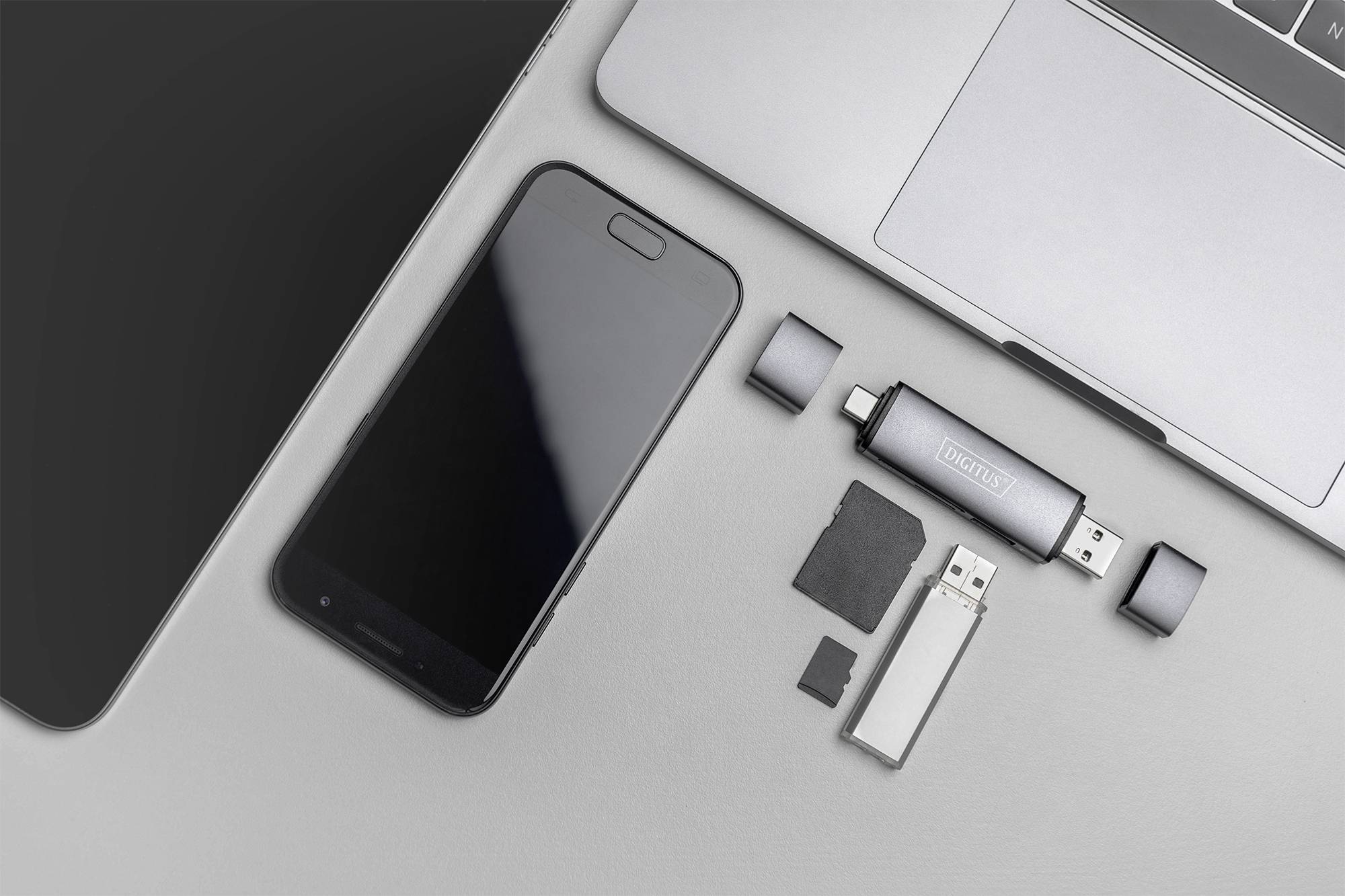 Smartphone, laptop, USB stick, microSD card and adapter on a grey surface. Technical devices for digital storage and connectivity tasks.