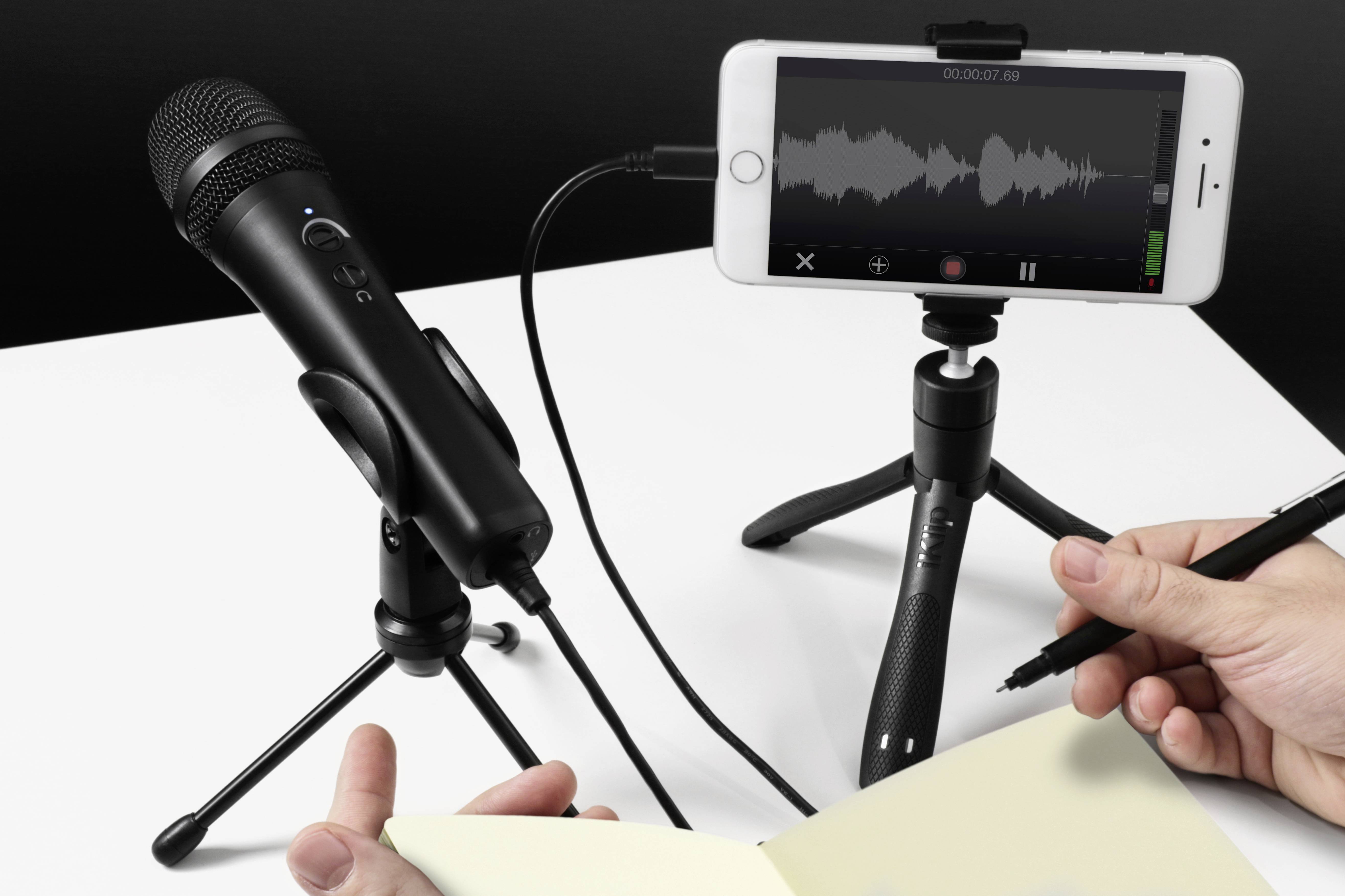 A microphone and a smartphone on tripods, connected by cable. An audio recording application is visible on the smartphone. One person is holding a pen and notebook.