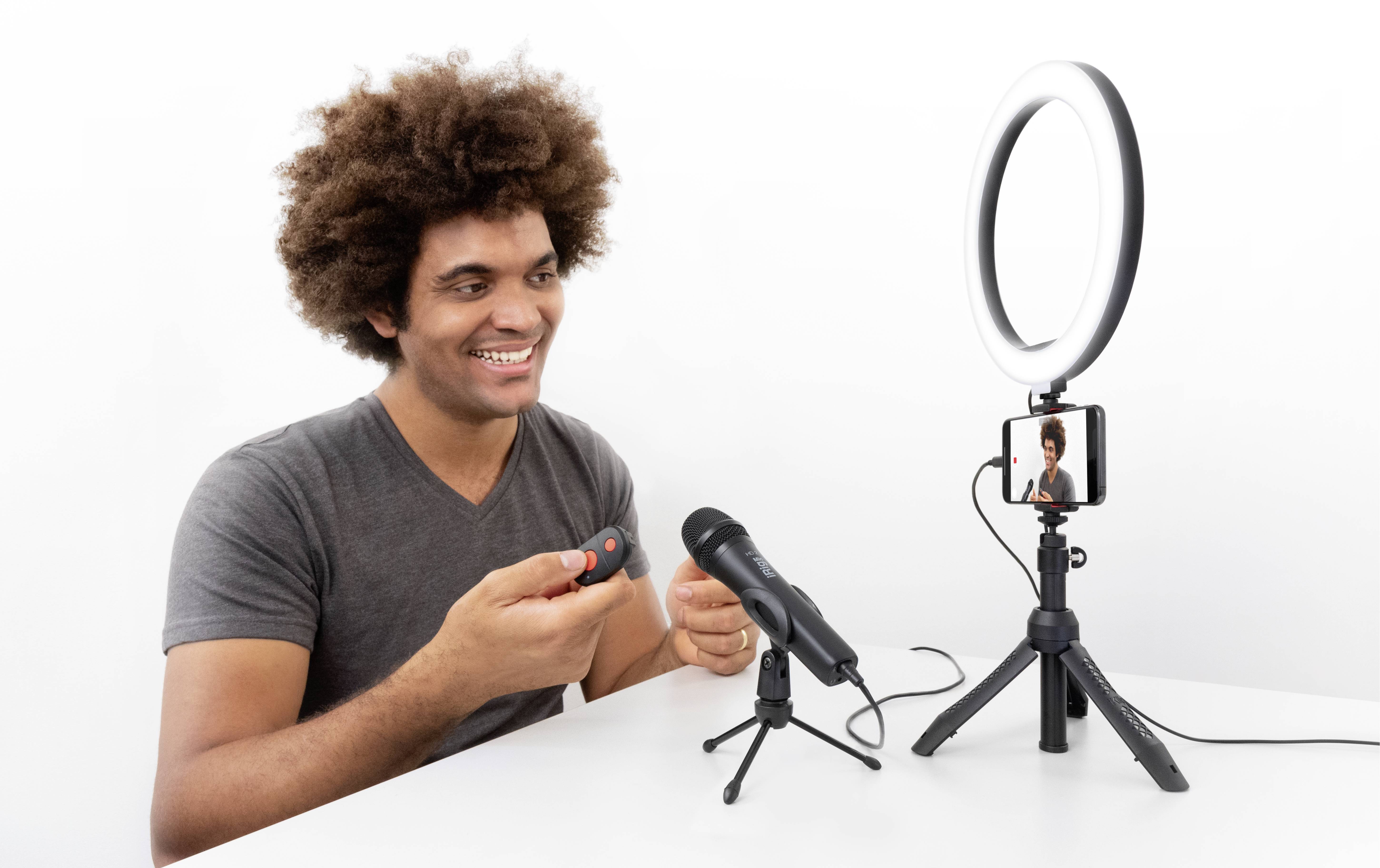 A man sits at a table with a microphone, a smartphone on a tripod and a ring light, while smiling and speaking.