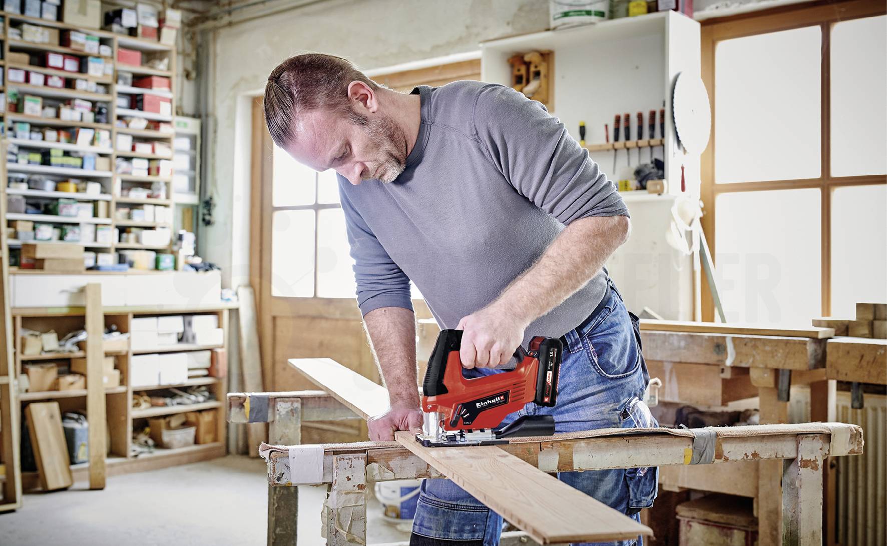 A man in a workshop is cutting a wooden board with a red jigsaw. In the background, shelves with tools and materials are visible.