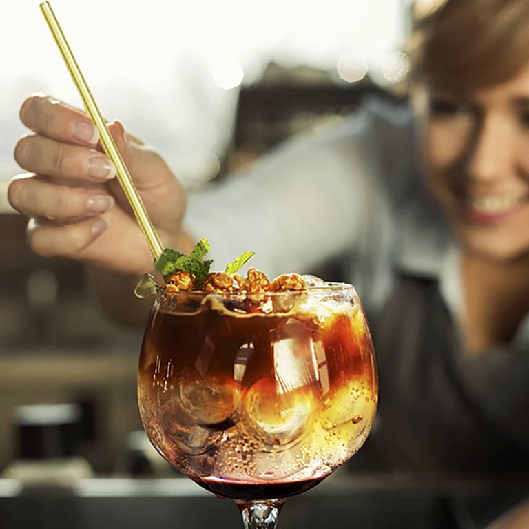 A person is holding a cocktail glass with ice and a golden straw, with a blurred background.