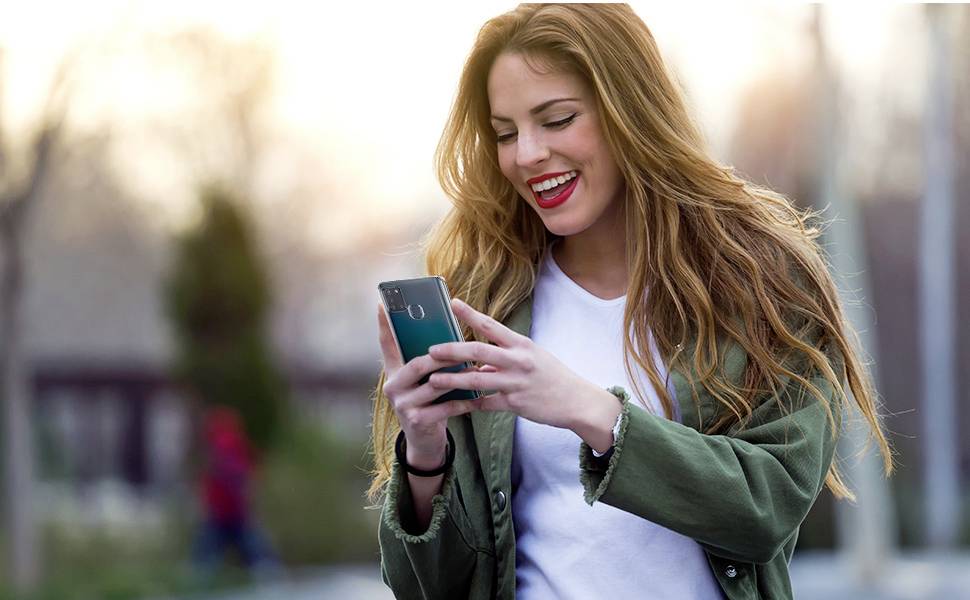 Woman with long hair smiles and looks at her smartphone outdoors.