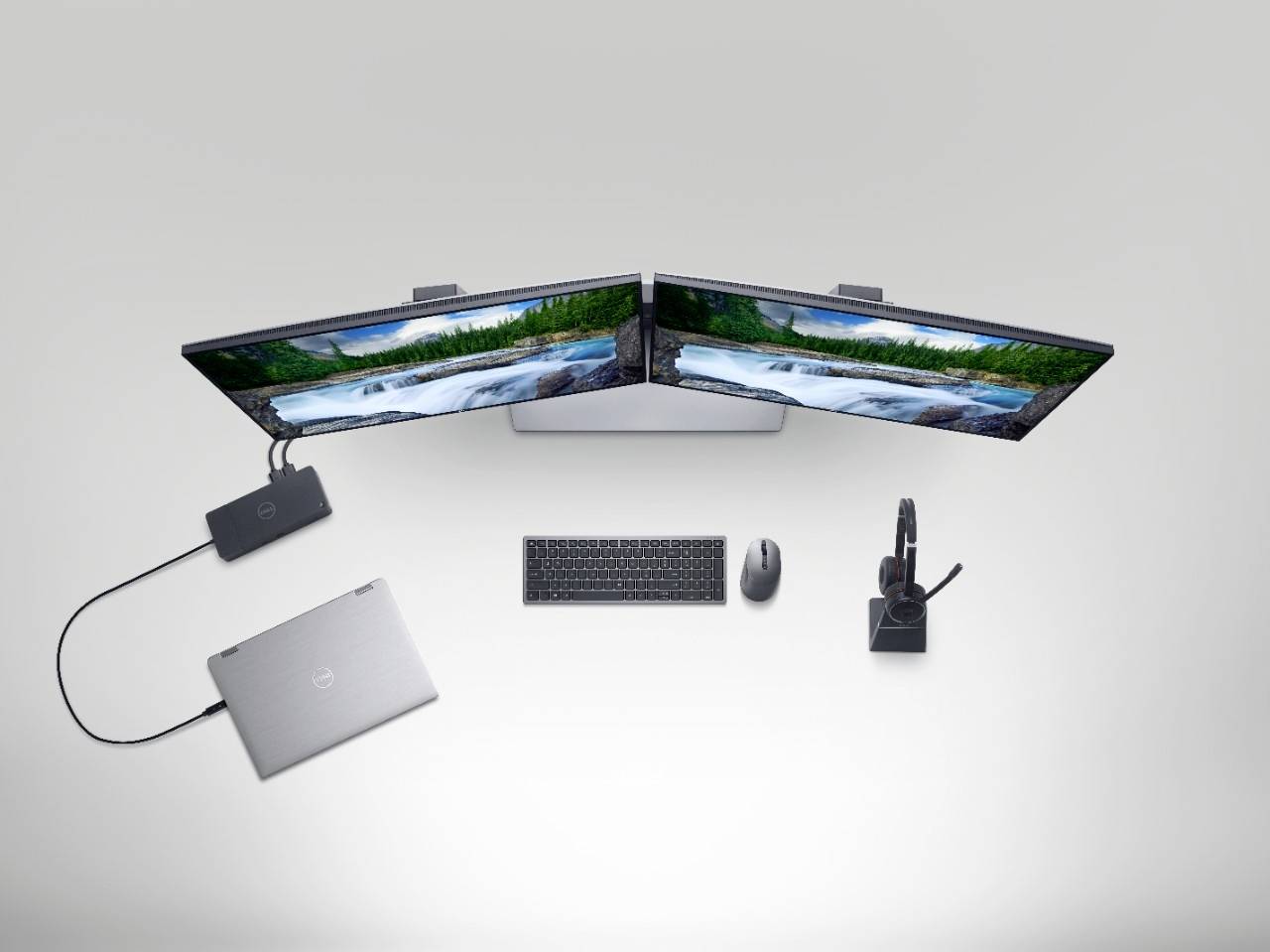 Two monitors are diagonally positioned, connected to a laptop. A keyboard and mouse are centrally placed, with headphones nearby.