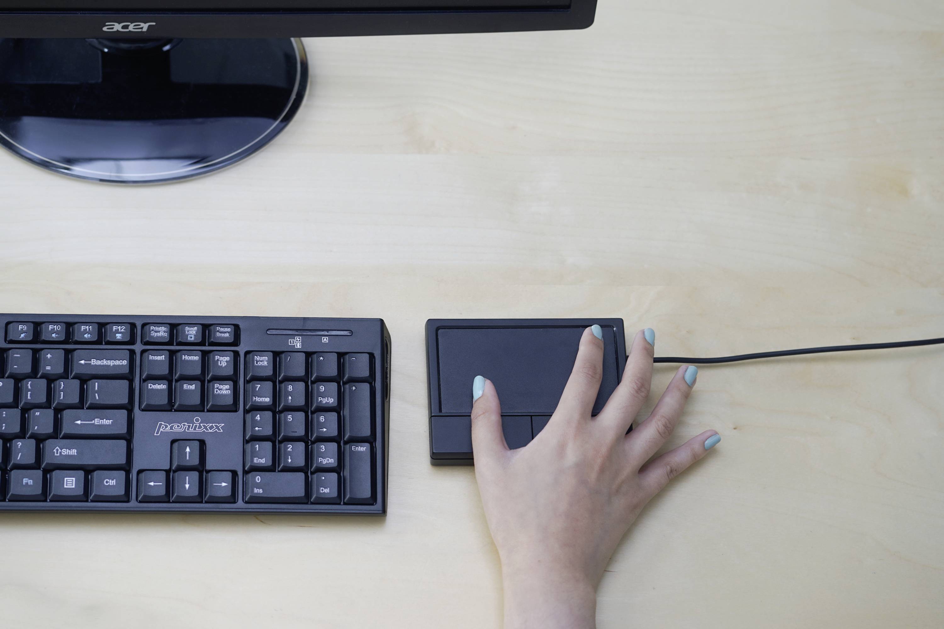 A hand is operating a touchpad next to a keyboard on a wooden table. A computer screen is partially visible at the top.