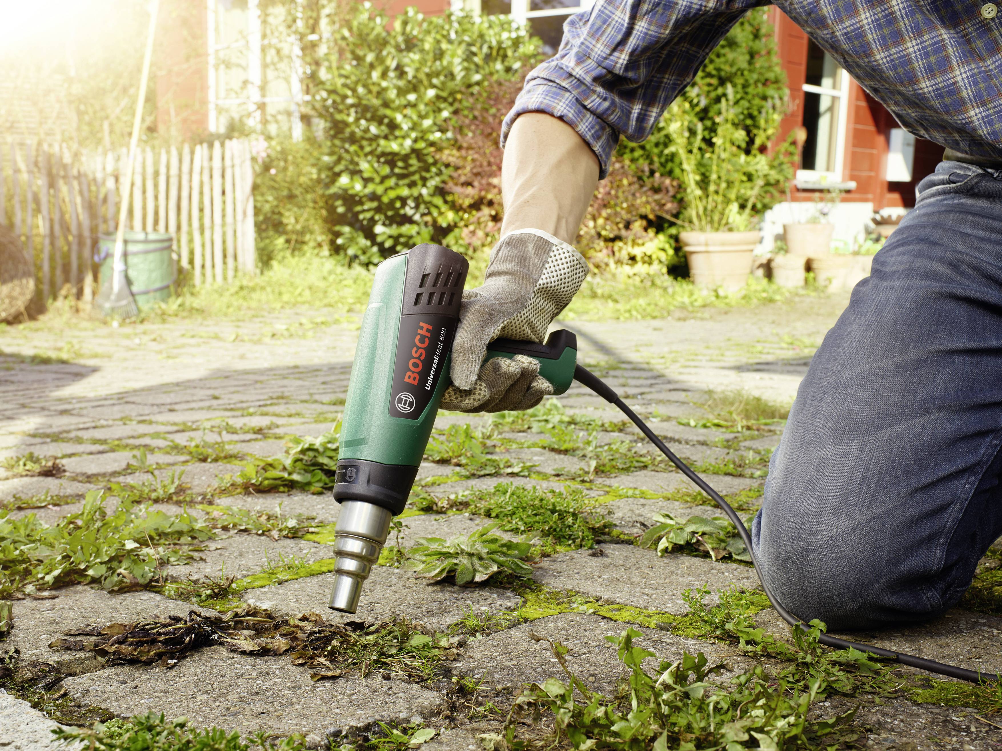 A person is using a hot air gun to remove weeds from a paved pathway. The background shows a house facade and garden.