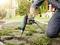 A person is using a hot air gun to remove weeds from a paved pathway. The background shows a house facade and garden.