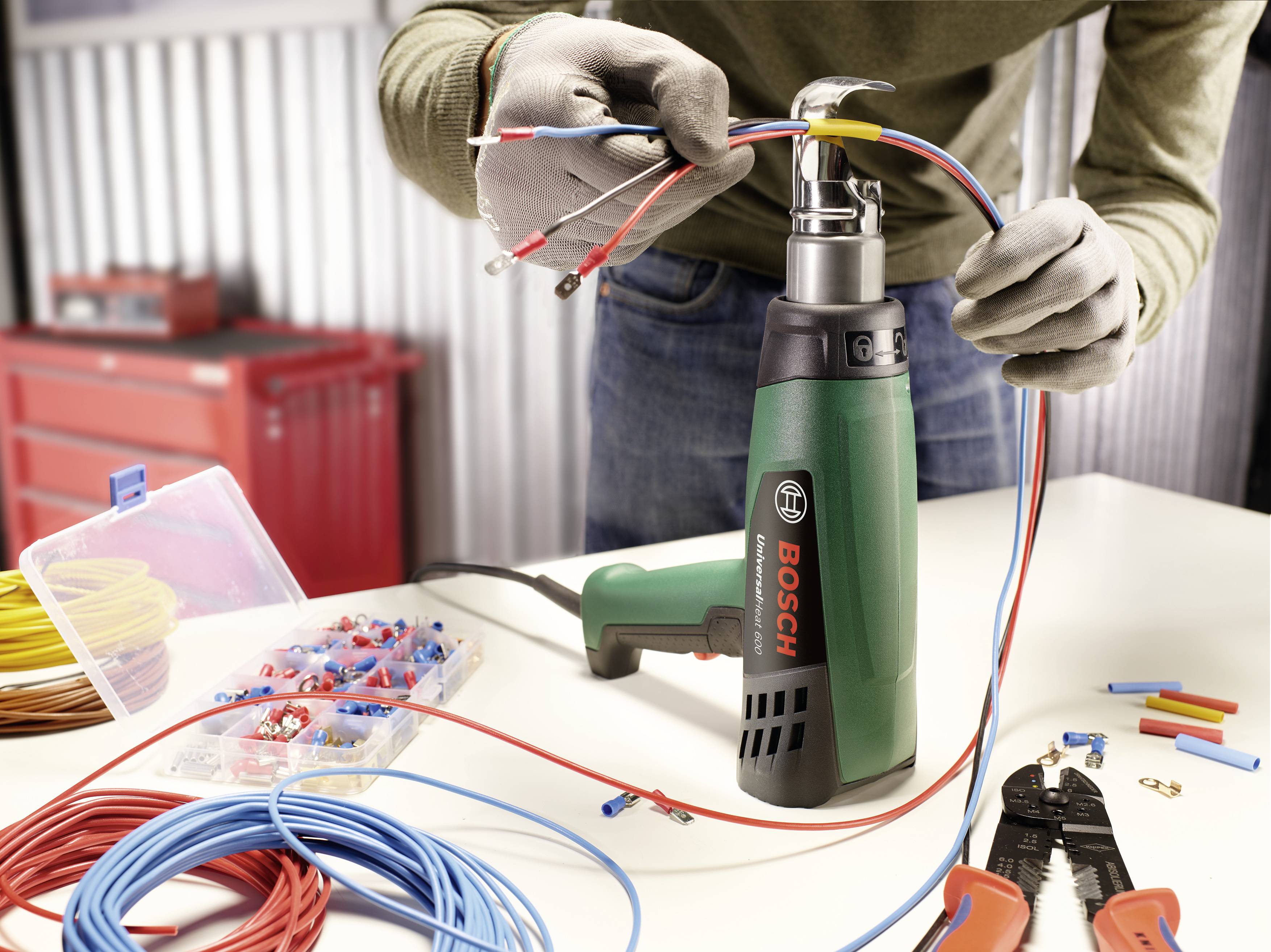 A person is working with a green Bosch heat gun and various cables on a table, surrounded by tools.