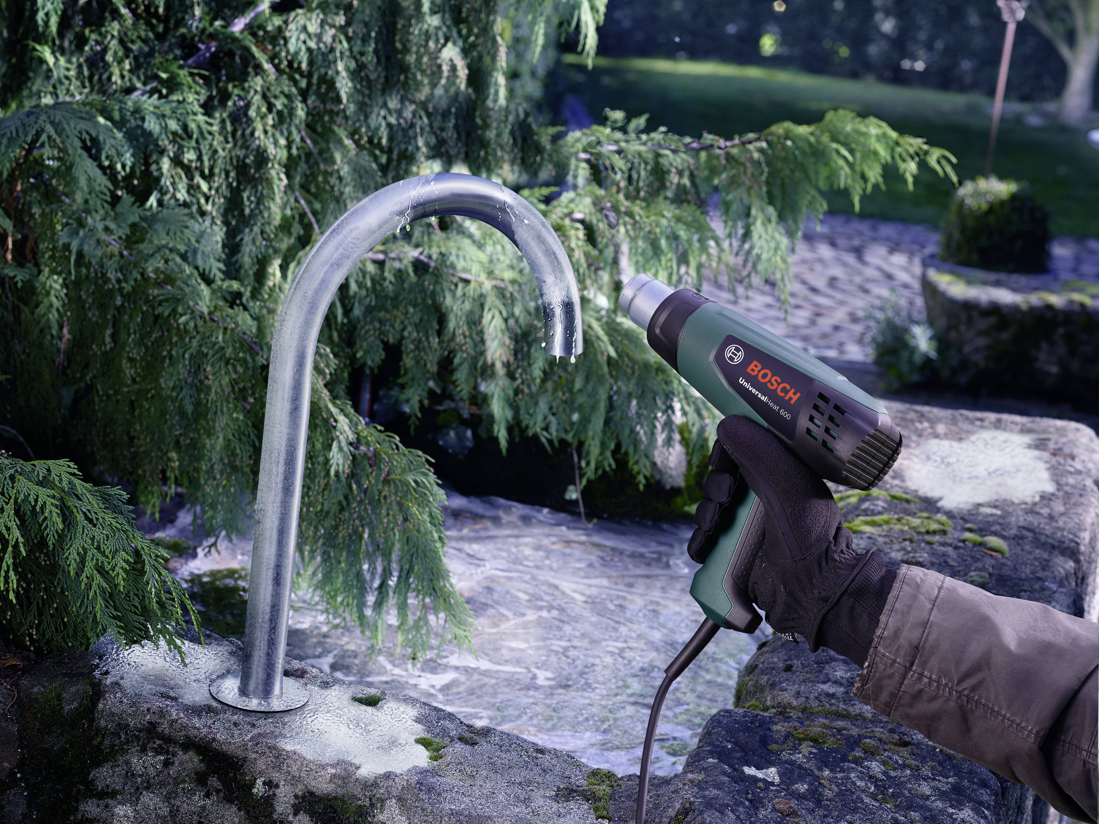A person is holding a hot air blower over a metal pipe outdoors. The background shows plants and a paved area.