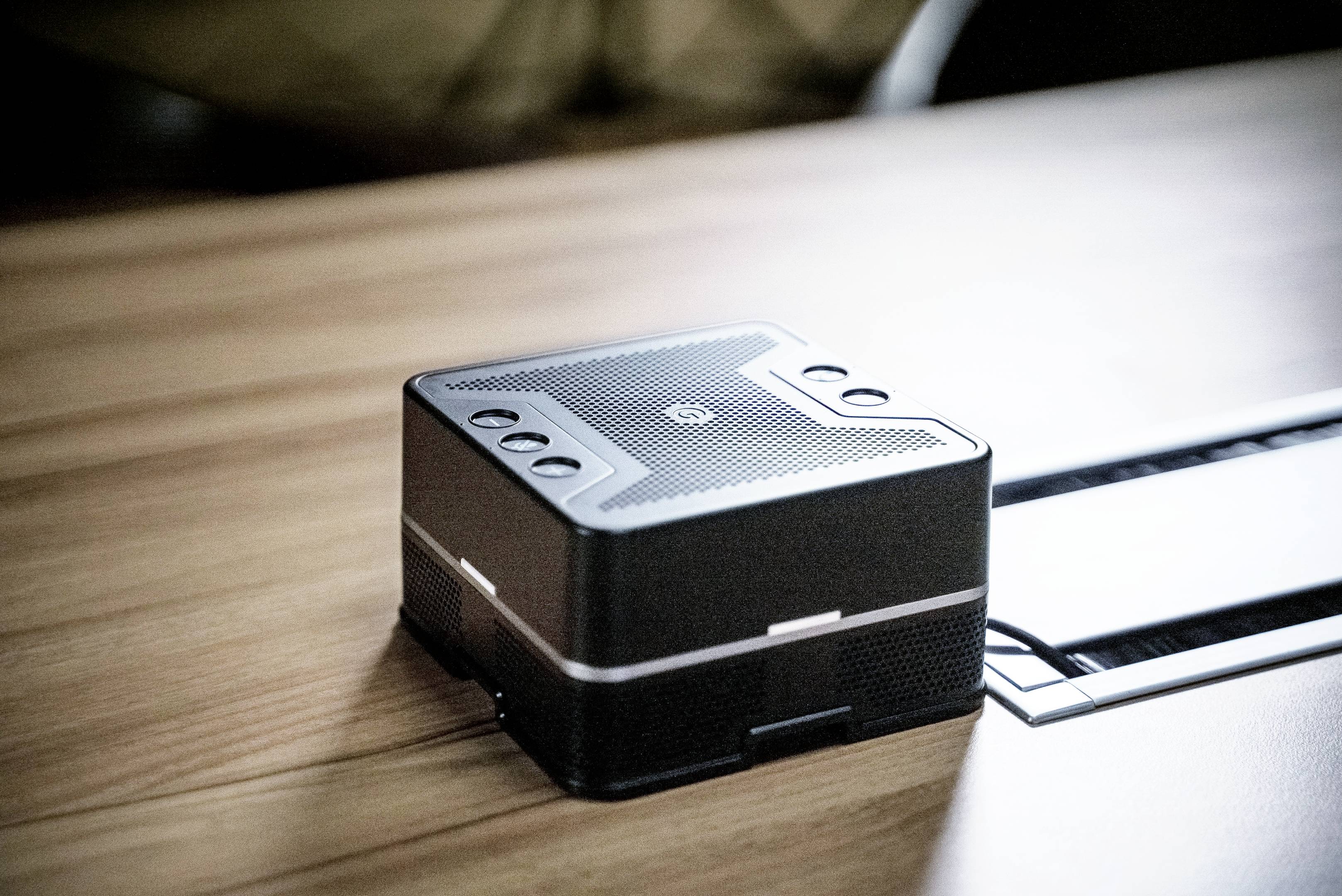 A black, square speaker with buttons on the top surface, sits on a wooden table.