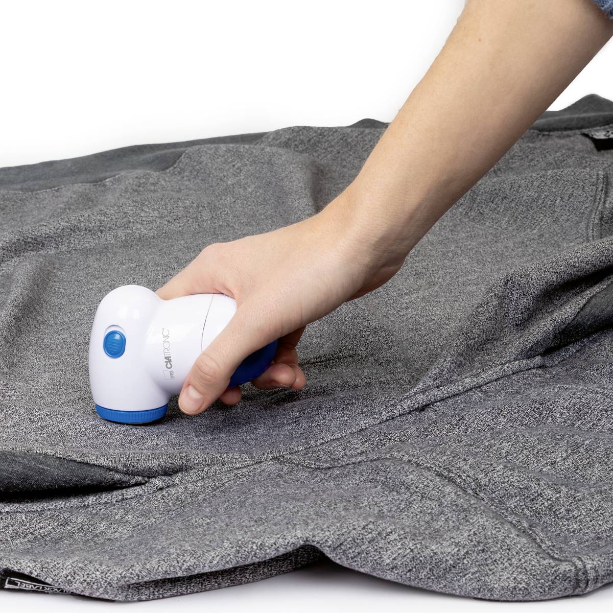 A hand is using an electric lint remover on a grey fabric to remove lint and fluff.